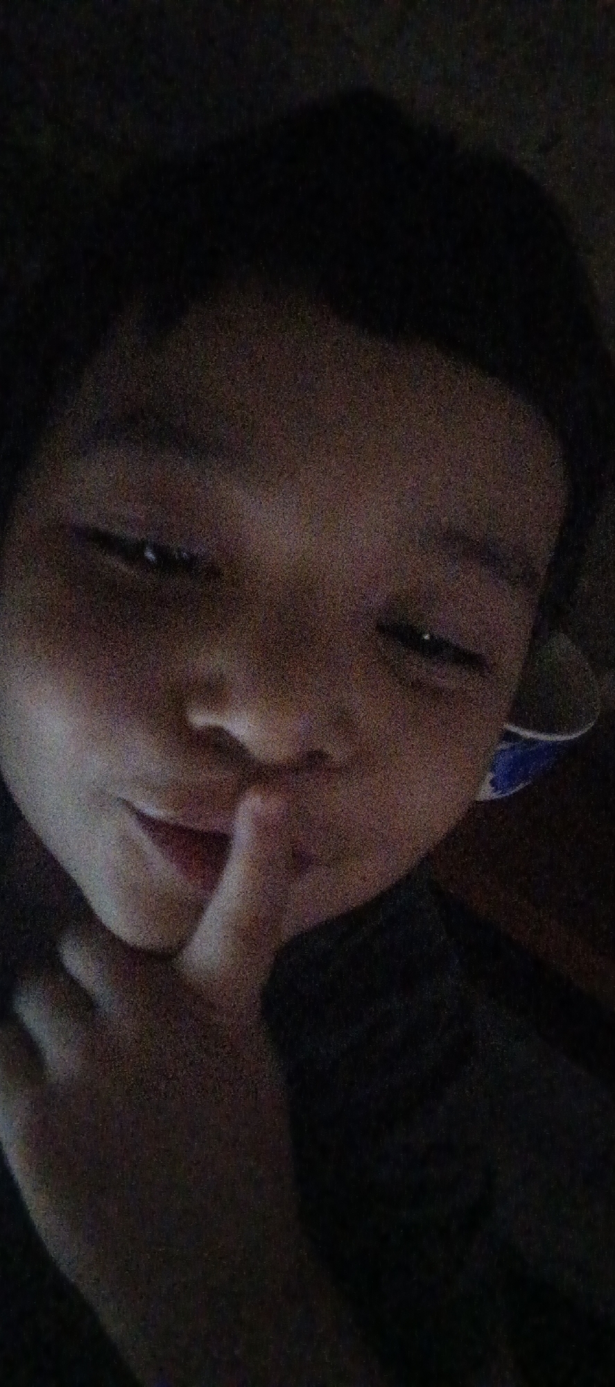 The image shows a close-up, low-light, grainy, forward-facing selfie of a young person's face. The individual has their index finger pressed against their lips, indicating a "shushing" gesture. Their eyes are partially visible, and their mouth is slightly open. The background is dark and blurred, with hints of what might be a dark garment or shadow. A portion of a blue and white patterned object, possibly a cup or bowl, is visible to the right of the person's head. The overall scene is dark and indistinct, with limited detail discernible due to the low lighting and image quality. There is no information within the image itself that would allow for the identification of the specific location in Shushary, Russia.