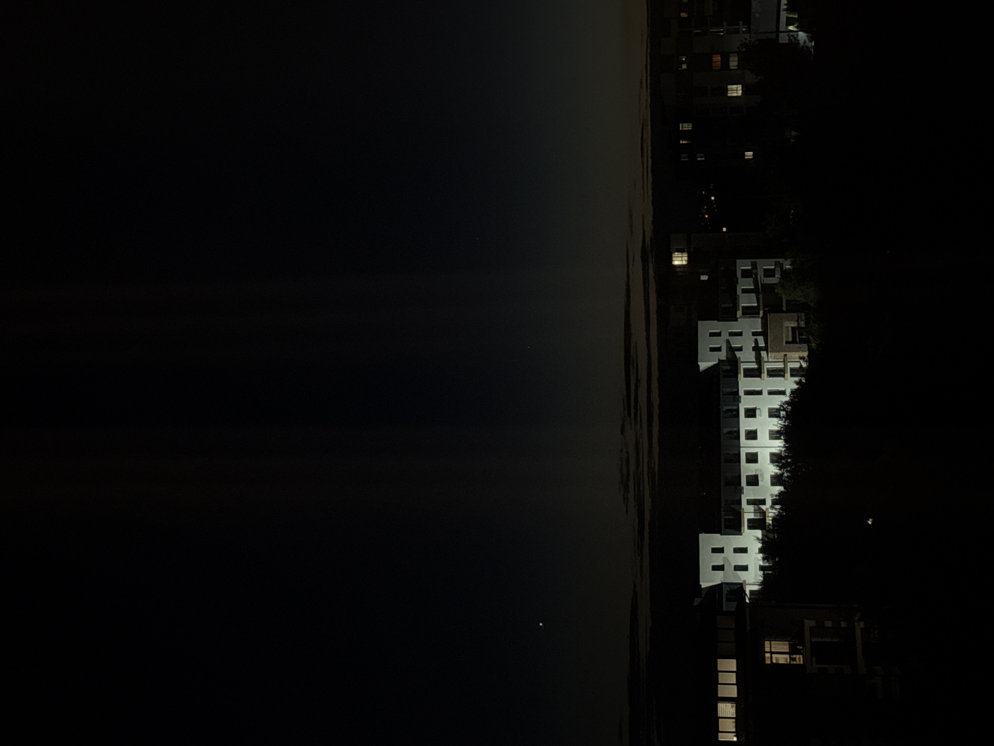 A long exposure night shot captures a city skyline at dusk. The sky is a deep, dark blue, transitioning to black at the top of the frame, with subtle, lighter grey bands of clouds near the horizon. A single, bright star or planet is visible in the upper left quadrant.
Below the sky, a low-lying building complex with several stories is illuminated by interior lights, creating a grid of warm, yellow and white squares against the dark exterior. The buildings appear to be modern, with a light-colored facade. Trees and foliage, silhouetted by the darkness, obscure the lower portions of the buildings.
In the far distance, beyond the main building complex, other structures are faintly visible, some with small, lit windows. The overall impression is one of a quiet, urban night scene.