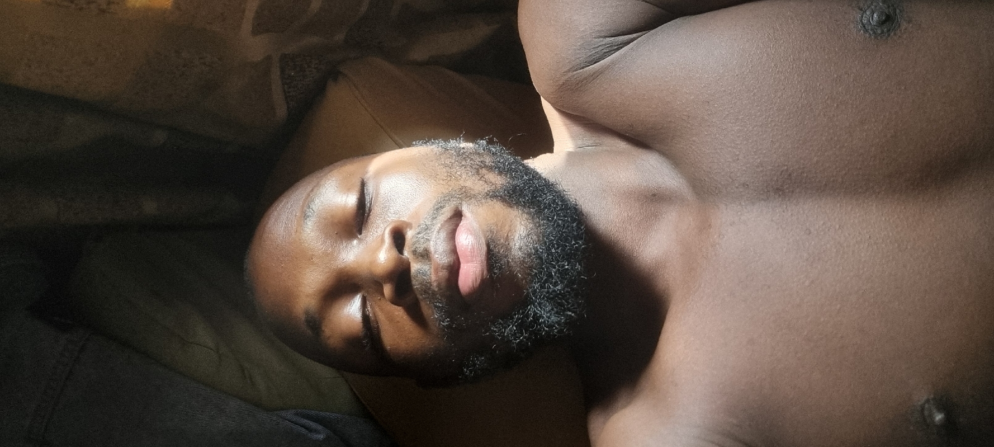 A shirtless, dark-skinned man with a beard is lying down. His eyes are closed, and his body is illuminated by sunlight. The background is blurred, but it appears to be a bed or couch. The location is Unknown, Unknown.