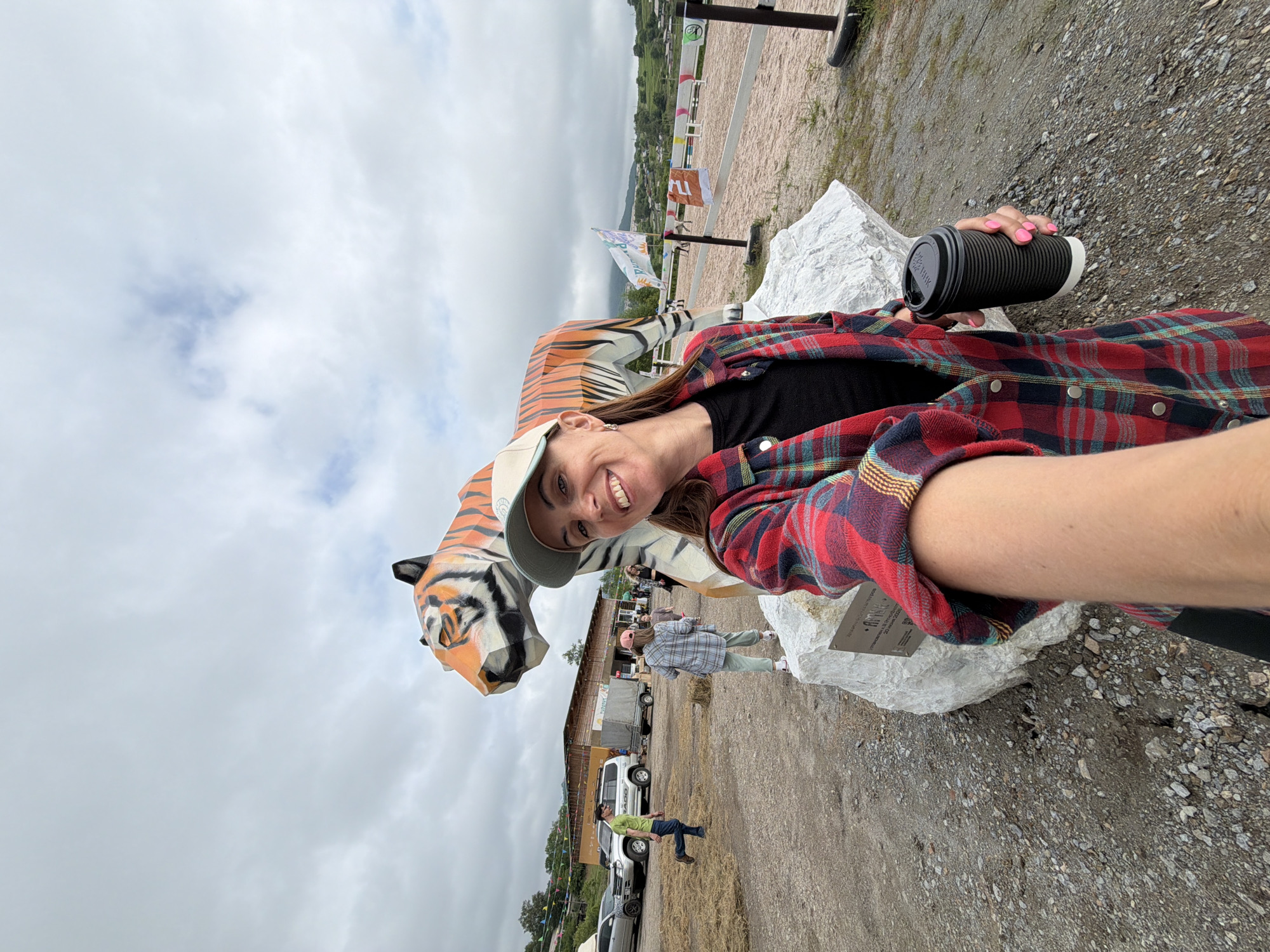 A woman takes a selfie in front of a large tiger sculpture in Теплоозёрск, Russia. She is wearing a red and blue plaid shirt, a black shirt underneath, and a light-colored baseball cap. She is holding a black coffee cup in her right hand, which has bright pink fingernails. The tiger sculpture is orange and black with striped patterns, and it is placed on a large white rock. Behind the sculpture, a dirt area is visible with several people walking around, some parked vehicles, and a building. Further in the background, there is a horse riding arena with flags and fences, and beyond that, rolling green hills with houses and trees under a cloudy sky. The foreground is dominated by the woman and the sculpture, with a gravelly ground surface.
