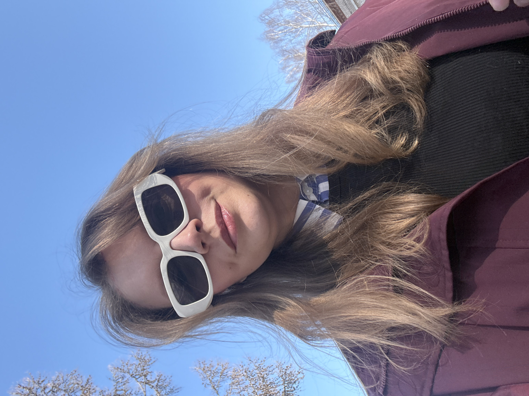 A young woman poses for a photo outdoors on a sunny day. She is wearing white-rimmed sunglasses, a black ribbed shirt, and a maroon jacket. A blue and white checkered shirt is partially visible under the black shirt. Her long, light brown hair is loosely styled. The background features a clear blue sky and bare tree branches, possibly indicating early spring. The location is not apparent from the provided image, so the location in the city of Izhevsk, Russia can not be determined.