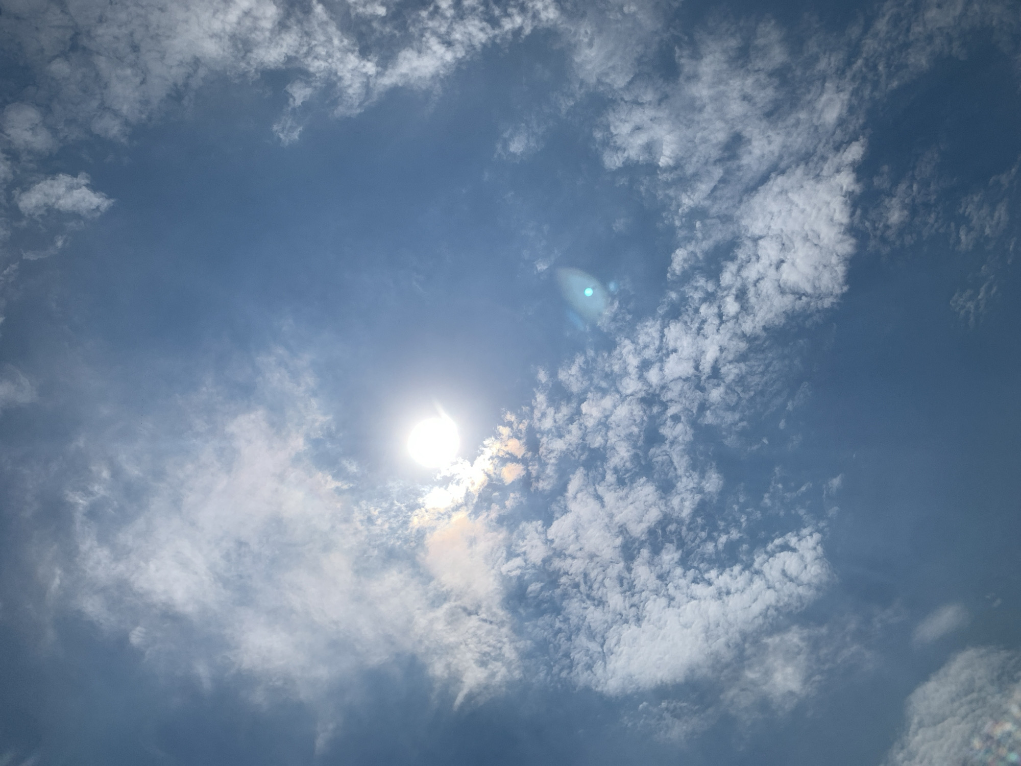 This is a photograph of a bright sun partially obscured by scattered white clouds against a clear blue sky. The sun is located in the upper left quadrant of the image, casting a strong glare. There are no people, animals, or man-made objects visible in the image. The scene appears to be an outdoor, daytime environment with a focus on the sky and atmospheric conditions. The location in Khazar Raion, Azerbaijan is not discernible from the visual information provided in the image.