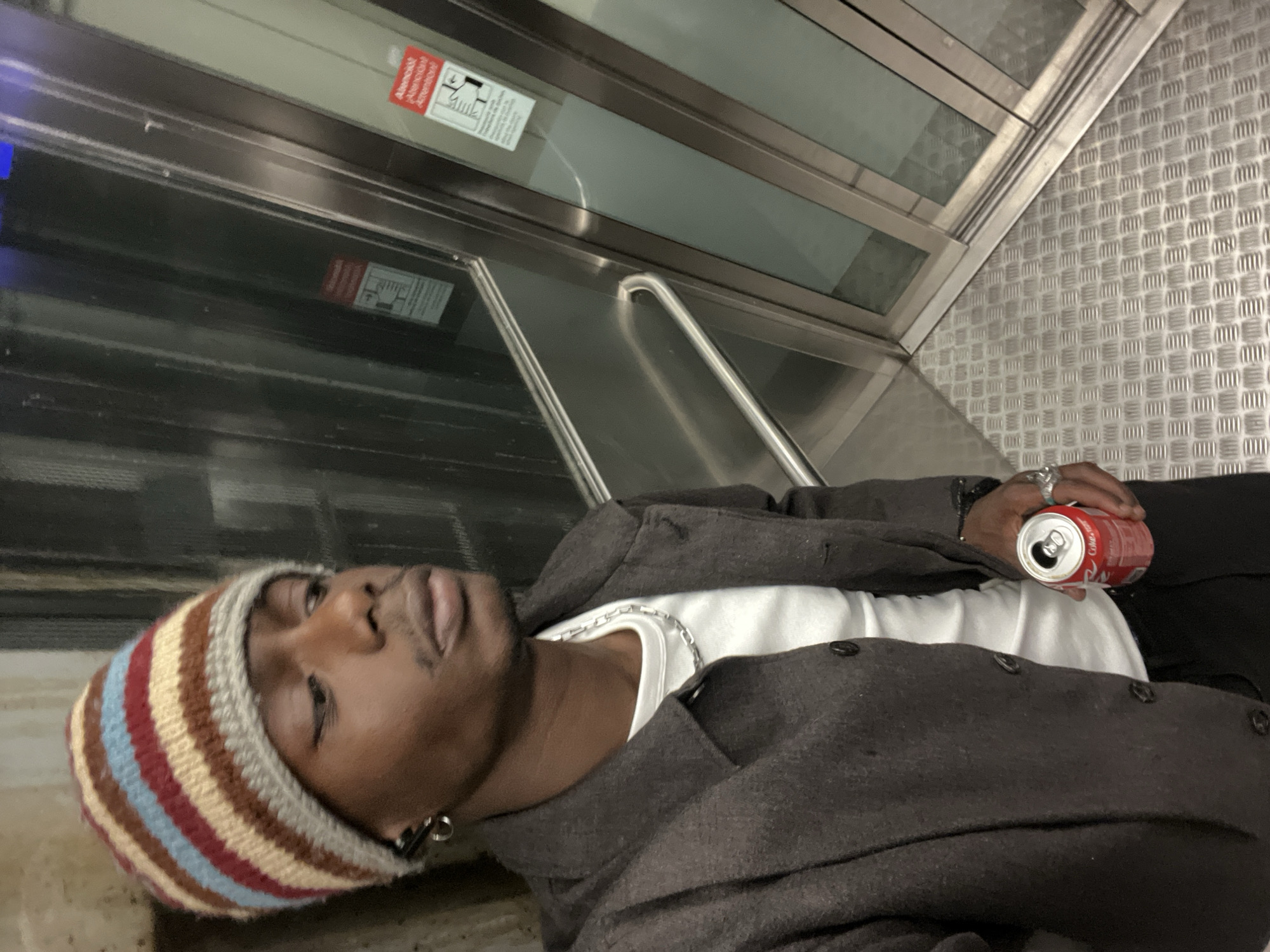 A young man with dark skin and a striped beanie is lying on the floor of what appears to be an elevator. He is wearing a white t-shirt, a silver chain, and a dark brown jacket. He is holding an open can of Coca-Cola in his right hand. The elevator has metal walls and a metal floor with a textured surface. There is a sign in the upper left corner of the frame. The photograph was taken in Barcelona, Spain.