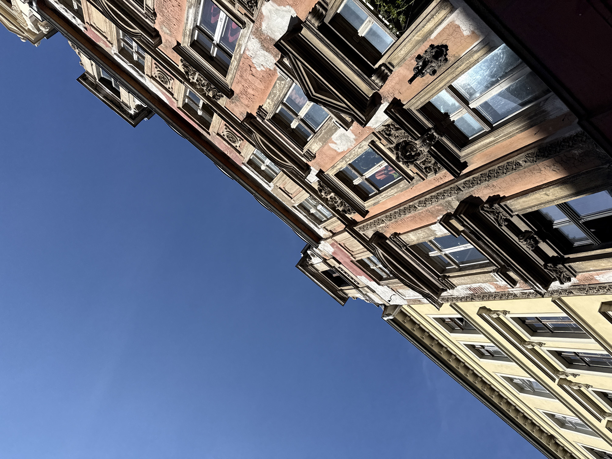 An upward angle shot shows two buildings in Görlitz, Germany, under a clear blue sky. The building on the right is a weathered, reddish-orange structure with elaborate architectural details around its windows, including ornate carvings and decorative trim. The plaster on this building is peeling in several places, revealing brickwork beneath. The building on the left is a lighter yellow, with simpler window frames and a smooth facade. Both buildings appear to be multi-story. The image captures the textures and colors of the aged architecture against the bright sky, suggesting a historic district of the city.