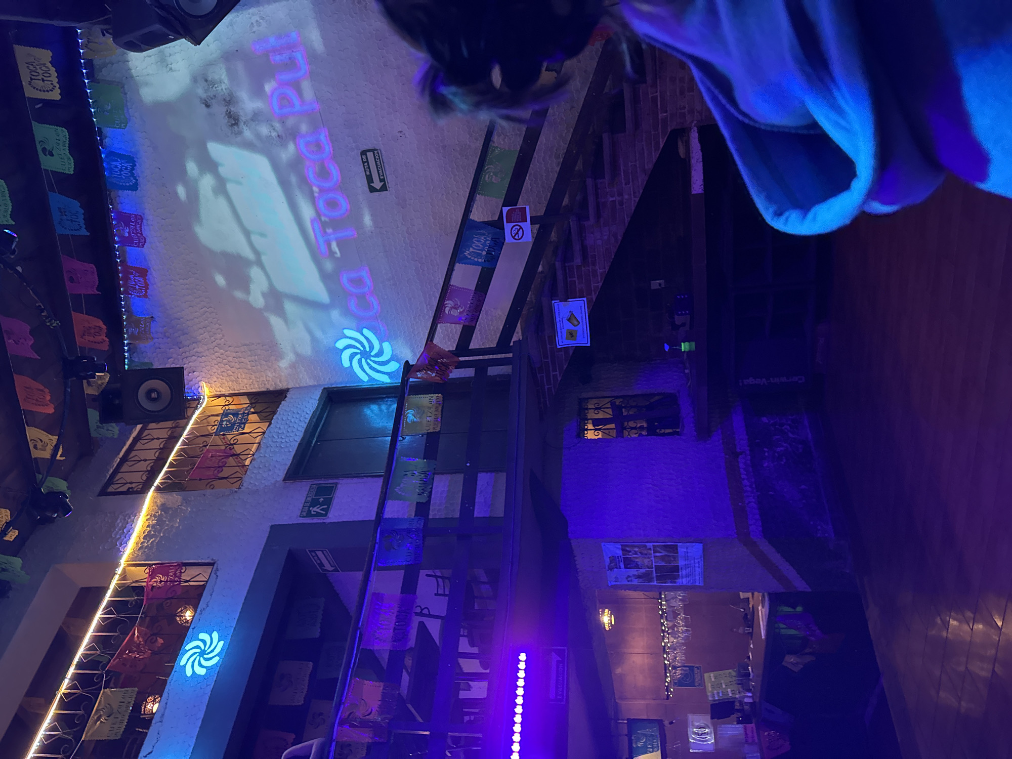The image shows an indoor shot of "Toca Toca Pulqueria" in Cuetzalan, Mexico. The interior is lit with blue and purple lights. Decorations include "papel picado" banners, some with the establishment's name. The name "Toca Toca Pul" is projected onto a wall. Speakers are mounted on the wall. A stairway is present on the right side of the image. Other signage includes "Ruta de Evacuación" and "Evacuación." A person partially occupies the upper center and right side of the image, obstructing the view.