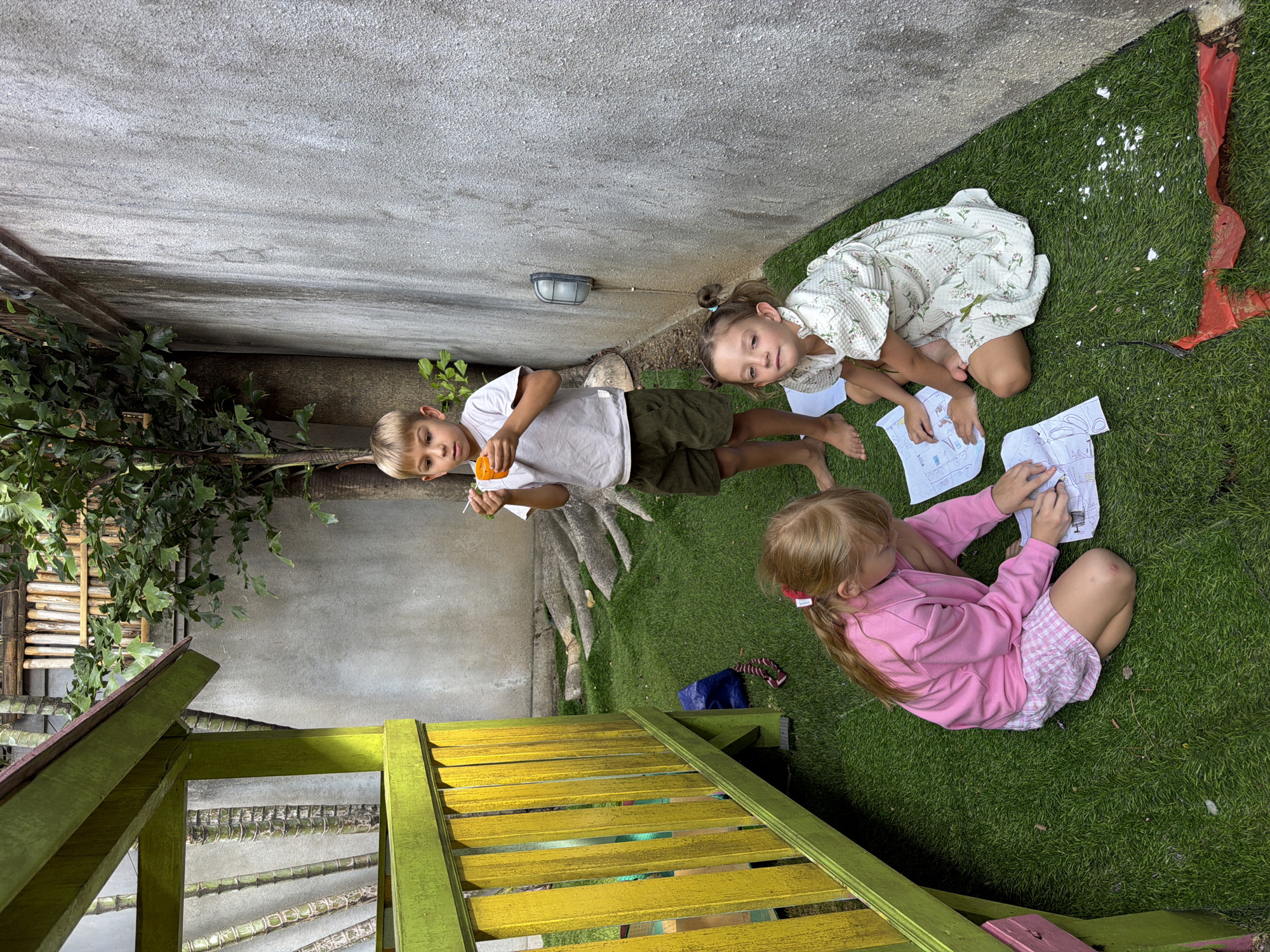 In Kuta, Indonesia, three children are engaged in an activity on a patch of artificial turf adjacent to a rough-textured concrete wall. A young boy, standing, holds an orange object and a white utensil, appearing to be eating or playing. In front of him, two girls are seated on the turf, engrossed in what seem to be papers laid out before them. One girl, wearing a pink top and a checkered skirt, is drawing or writing. The other girl, in a floral-patterned dress, is also looking down at her paper. To the left, a brightly painted yellow and green structure, possibly part of a playhouse, occupies a significant portion of the frame. The environment suggests an outdoor recreational area within an urban setting.