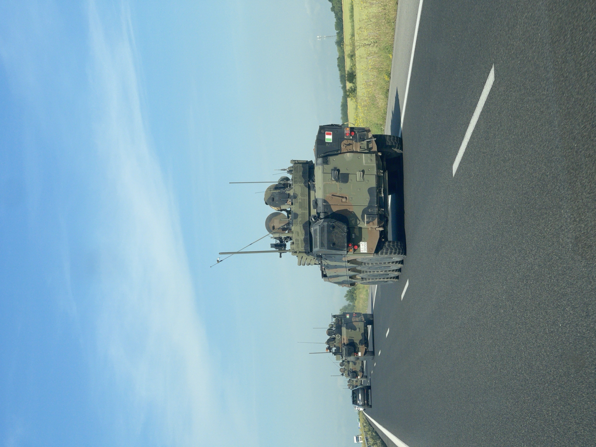 A military convoy consisting of several armored personnel carriers or infantry fighting vehicles is depicted traveling along a multi-lane highway under a clear blue sky. The image is taken from a following vehicle, showing the convoy stretching into the distance. The military vehicles are painted in camouflage patterns, primarily green and brown.
The lead vehicle in the convoy, closest to the viewer, is a distinctly Italian military vehicle. It features a vertical red, white, and green Italian flag prominently displayed on its rear. Below the flag, an identification plate bearing the text "EI 2039" is visible, with "EI" commonly denoting "Esercito Italiano" (Italian Army). Further back in the convoy, another vehicle displays a horizontal red, white, and green flag on its rear, consistent with the flag of Hungary.
The road is paved with asphalt, featuring clear white dashed lane markings and a solid white line along the shoulder. To the right of the highway, a verdant landscape of fields and scattered trees extends under the bright sky, with a power line tower visible on the horizon. The time of day is daytime, characterized by bright sunlight and excellent visibility. The setting in Загорци, Bulgaria, combined with the presence of Italian and Hungarian military vehicles, suggests a multinational military operation, possibly a NATO exercise or troop movement.