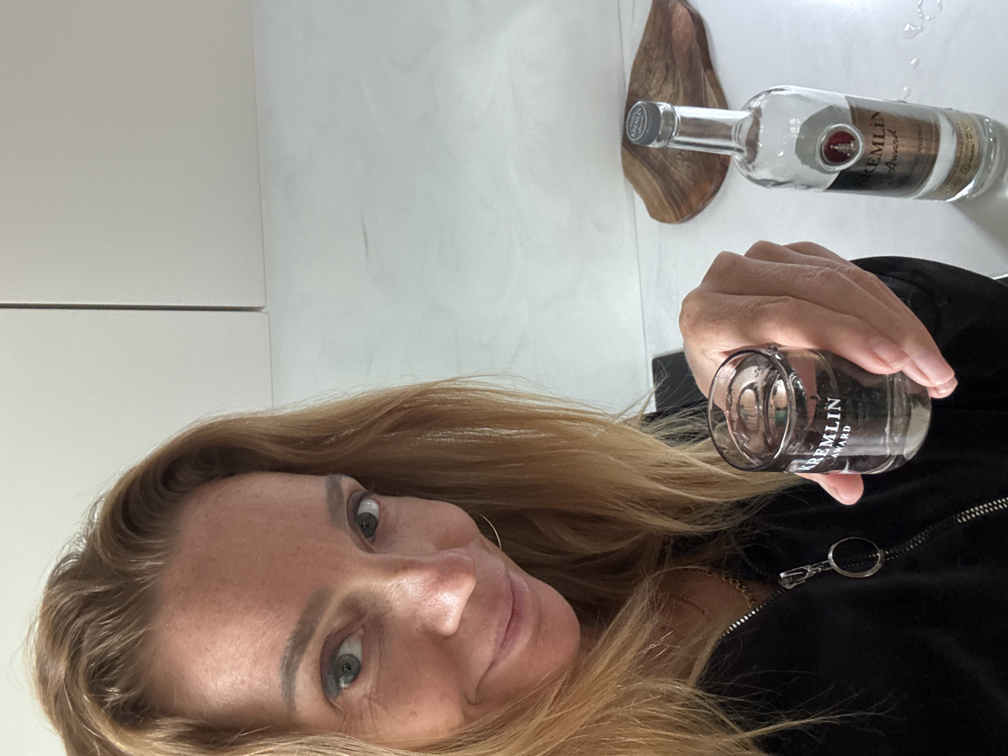 A blonde-haired woman holds a glass of Kremlin Award vodka, appearing to pose for a selfie. A bottle of the same vodka sits on a white surface, partially on a wooden object. The surface resembles a table or countertop, with a possible wall in the background. The environment suggests an indoor setting, likely a home or apartment. The city of Steklyannyi, Russia, is not directly evident in the image, but the vodka brand provides an indirect connection to the country.