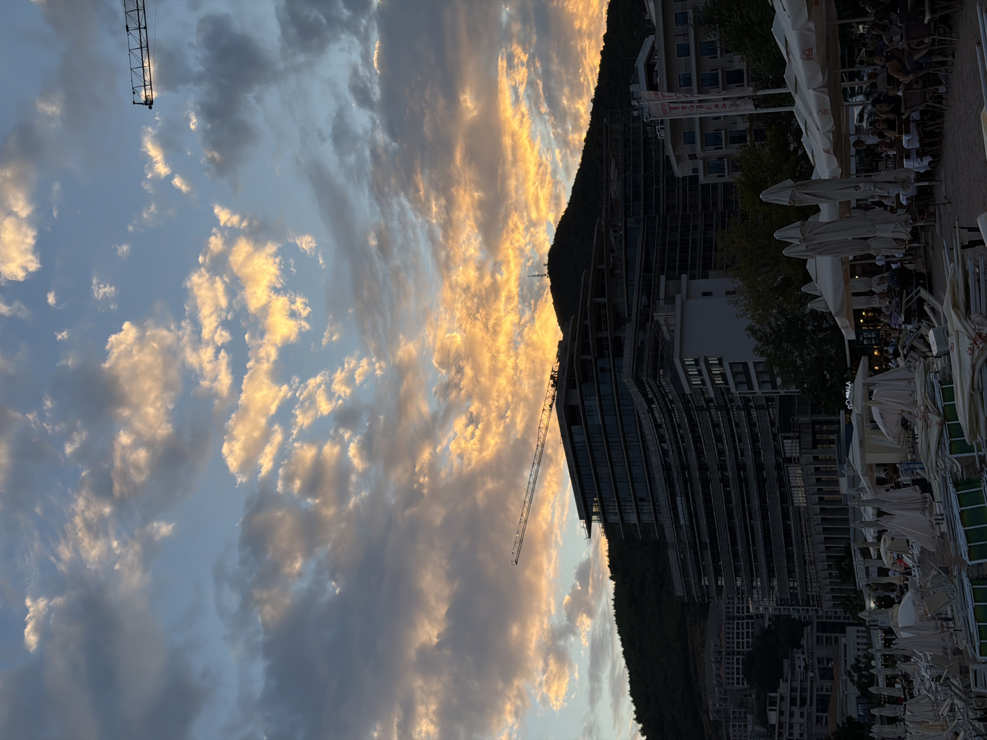 An outdoor, eye-level, full shot captures a coastal city scene in Budva, Montenegro, under a dramatic sunset sky. The sky is filled with voluminous clouds, illuminated from behind by the setting sun, casting vibrant hues of orange, yellow, and pink. A construction crane is visible in the upper right corner, and another crane is seen extending horizontally across the mid-ground.
Dominating the mid-ground is a large, modern, multi-story building under construction, its dark facade contrasting with the bright sky. The building's architecture features a stepped, terraced design with numerous balconies and large windows. Behind this structure, a dark, tree-covered hillside rises. To the right of the unfinished building, a completed, modern hotel building is visible, adorned with balconies and windows.
In the foreground, a beach area is set up with rows of tightly packed, closed white umbrellas and lounge chairs, indicating a public beach or resort area. A few people are scattered amongst the beach furniture, and some are seated at tables under open umbrellas in a section of the beach that appears to be a cafe or restaurant area. The overall impression is one of a developing coastal resort town at dusk, with a blend of ongoing construction and established tourist amenities.