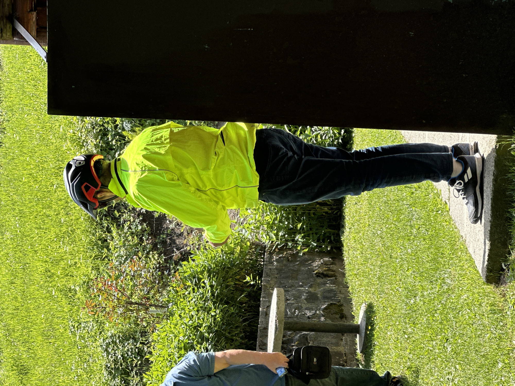 The image depicts two individuals outdoors, likely in a park or garden setting, with a backdrop of lush green grass and foliage. The primary subject, positioned on the right, is a person wearing a bright yellow, long-sleeved jacket and dark blue jeans, along with a black and red cycling helmet. This individual is standing next to a dark, vertical structure, partially obscuring their right side. Their stance suggests they are facing away from the camera, looking towards the left.
To the left of the frame, another person is visible from the waist down, wearing a blue shirt and dark jeans. They are holding a black messenger bag in their left hand and appear to be standing near a round, concrete table supported by a stone base. This individual is facing towards the right.
The environment suggests a sunny day, with dappled light illuminating the grassy areas and plants. The presence of the cycling helmet on the main subject could indicate an activity related to cycling. The concrete table and stone wall suggest a possibly maintained outdoor area. The overall scene conveys a casual, outdoor gathering or moment. The specific location within Bare Kraljske, Montenegro, is not explicitly discernible from the visual elements present.
