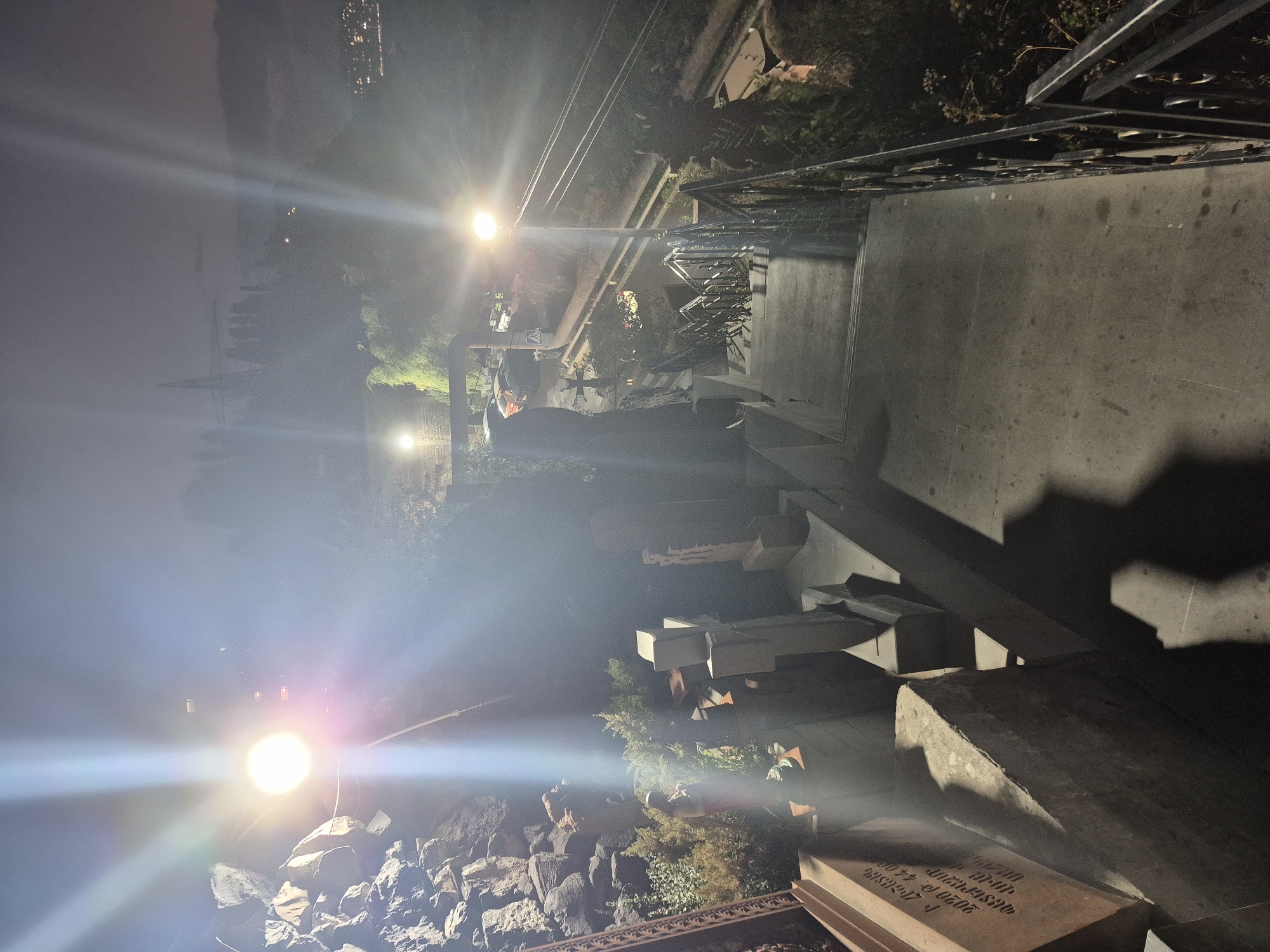The image captures a view from an elevated position looking down a stone staircase at night in Yerevan, Armenia. The staircase, flanked by a wrought-iron railing on the right, leads down into a dimly lit area. On the left side of the staircase, a collection of gravestones and what appears to be a statue are visible, suggesting the location is a cemetery or memorial site. Large rocks are piled up to the left of the gravestones. Streetlights illuminate the scene, casting bright beams and creating lens flares that partially obscure details. In the background, trees and distant city lights are faintly visible under a dark sky, with a power line tower also present. The overall atmosphere is somber and illuminated by artificial light.