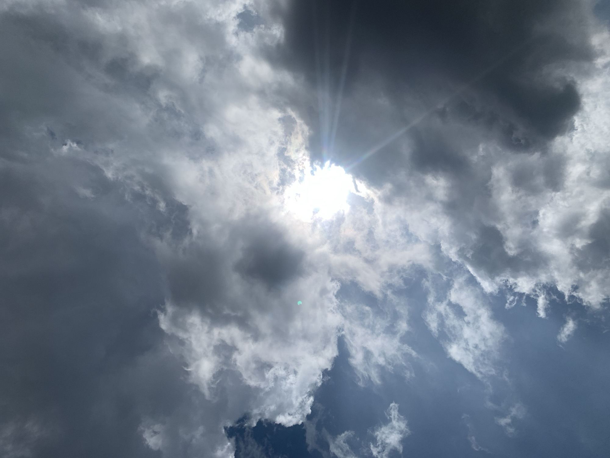The image shows a dramatic sky filled with dark, heavy clouds and rays of sunlight breaking through. The sun is a bright, almost blinding orb peeking from behind a dense cloud formation, creating visible sunbeams that fan outwards. The clouds vary in texture and color, ranging from deep gray to bright white where they are illuminated by the sun. There is a hint of blue sky visible in the lower right portion of the image, suggesting that the clouds are not completely covering the sky. The overall impression is one of a dynamic and possibly stormy sky. The scene is an outdoor sky view, likely captured during the daytime given the strong sunlight. No people, objects, or specific location cues are visible, beyond the sky itself. The weather appears to be overcast with sun breaking through, possibly indicating an approaching storm or the end of one. There is no visible text.