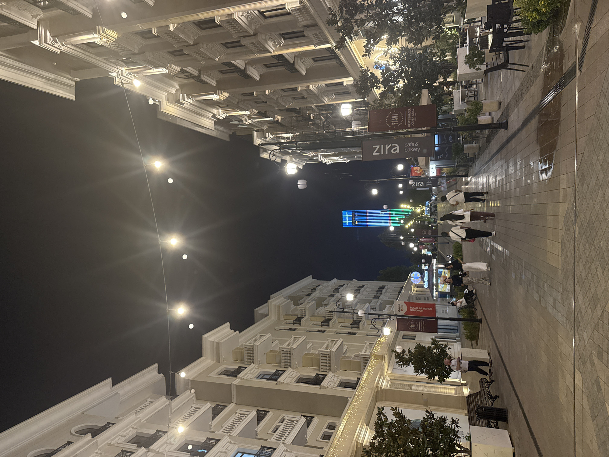 This image depicts a nighttime scene on a pedestrian street in Tashkent, Uzbekistan, likely part of the Sergeli district or a similar modern development. The street is lined with elegant, multi-story buildings with ornate architectural details, illuminated by warm exterior lights and strings of decorative bulbs overhead.
Several people are visible walking along the wide, paved street. On the left, a man in a white shirt and dark pants stands near a bench and a planter. Further down the street, a group of four individuals are walking away from the camera. On the right side of the street, several outdoor seating areas for cafes and restaurants are set up, with tables and chairs illuminated by streetlights.
In the distance, a tall, modern building is prominently lit with vertical bands of green and blue lights. Banners with text such as "Zira cafe & bakery" and "GOSHT MEAT & BURGER JOSPER GRILL" are displayed on lampposts along the street, indicating commercial establishments. The pavement appears slightly wet in places, suggesting recent rain. The overall atmosphere is one of a lively, well-lit urban environment at night.