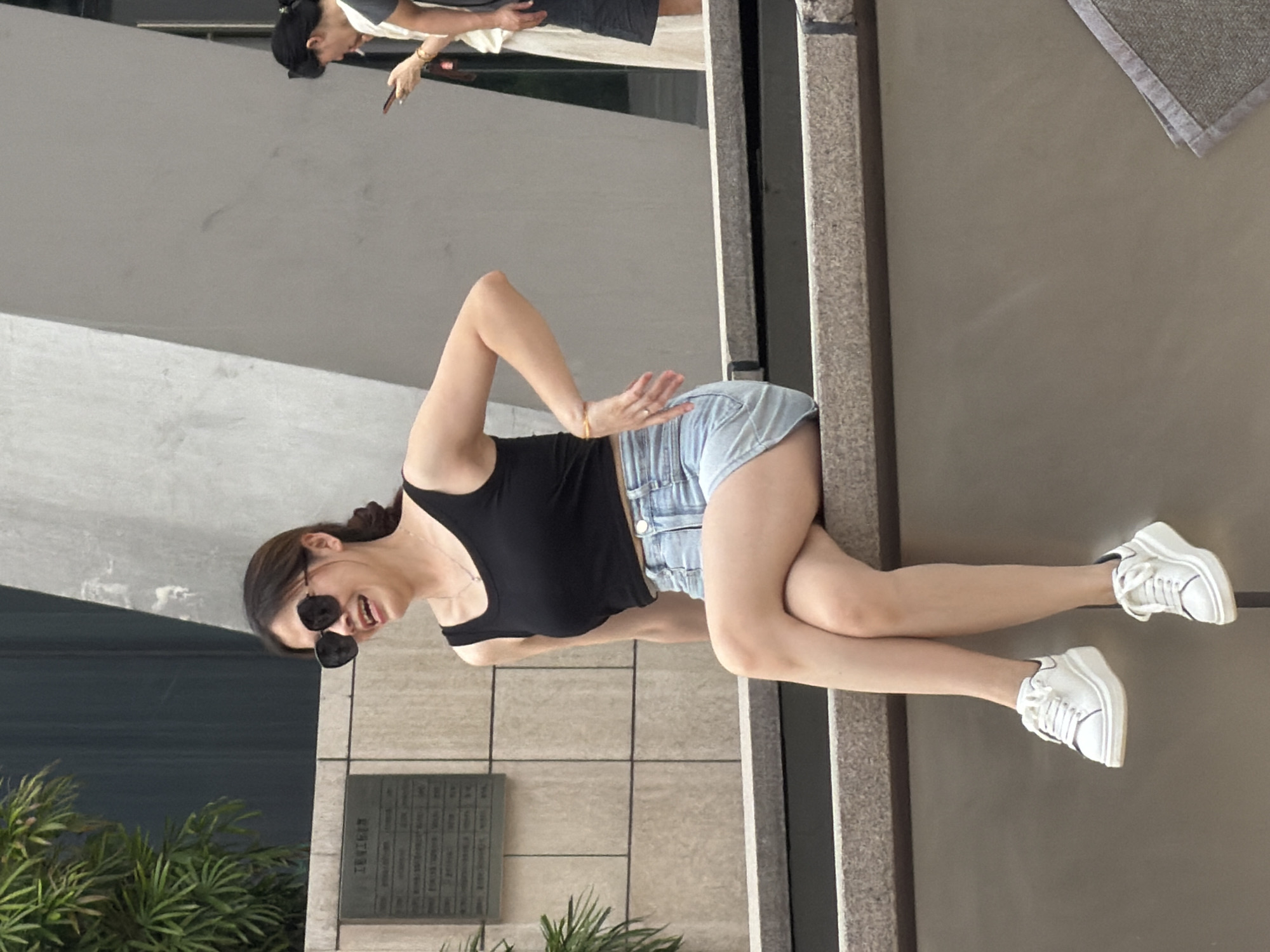 In Yuzhong District, China, a young Asian woman is sitting on a stone ledge outdoors, with her legs crossed and her feet resting in white sneakers. She is wearing a black tank top and light blue denim shorts, and large black sunglasses. Her right hand is resting on her hip. Her mouth is open as if she is laughing or speaking. To the right of the main subject, two other individuals are visible in the background. One person, dressed in a light-colored garment, is holding a smartphone. The other person, partially visible, is wearing a dark garment. The environment features a stone wall with a plaque on it and some greenery in the background.