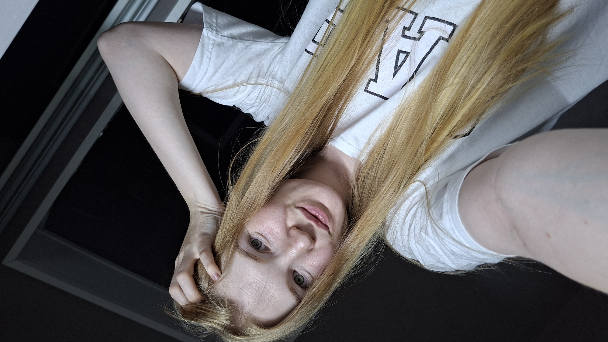 A young Caucasian woman with long, blonde hair and green eyes is captured in a close-up, high-angle selfie. She is wearing a white t-shirt with a large black graphic of the letter "A". Her left arm is bent, with her hand touching the top of her head, and her right arm is extended towards the camera, likely holding it. She has fair skin with light freckles across her nose and cheeks, and her lips are slightly parted as she smiles. The background is dark and appears to be an indoor setting, possibly near a window with a dark frame. The overall lighting is soft, highlighting her facial features. The location is identified as Chaikovsky, Russia.