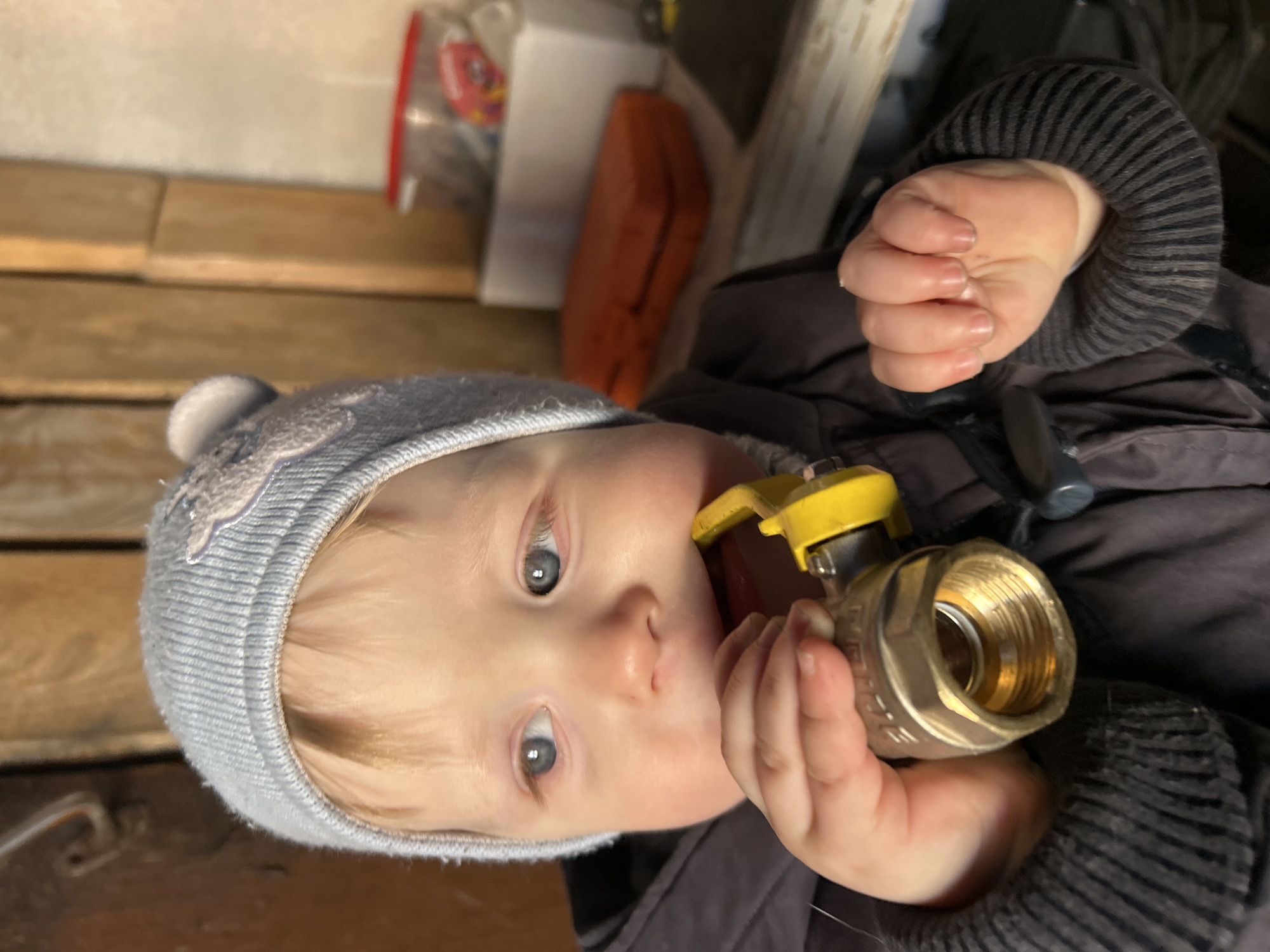 A young child with fair skin and blonde hair is holding a brass ball valve with a yellow handle to their mouth. The child is wearing a grey knit hat and a dark-colored jacket with a striped sleeve. The background consists of wooden planks and various items, including a white box, a clear plastic bag with red contents, and an orange case. The scene appears to be indoors, potentially a workshop or storage area. No information in this image indicates that the location is the city of Chaikovsky, Russia.