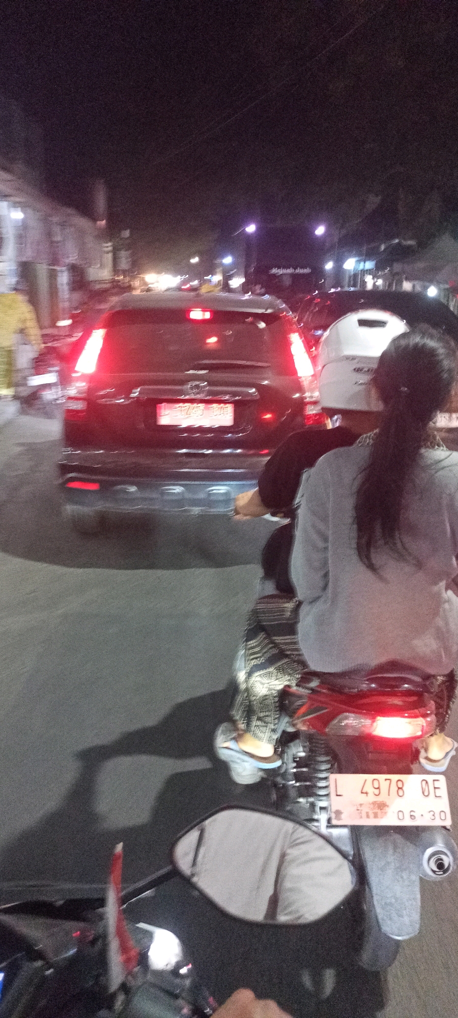 A nighttime, eye-level, medium shot captures a busy street scene in Singogalih, Indonesia. In the foreground, the back of a motorcycle is visible, with a license plate displaying "L 4978 OE 06.30". Two individuals are seated on the motorcycle; the person in the back is wearing a light gray long-sleeved shirt and a patterned skirt, with dark hair pulled back in a ponytail. A white helmet is on their head. A person in the front, partially obscured, is wearing a dark shirt and a white helmet. The motorcycle is moving on a paved road. Behind the motorcycle, a dark SUV with illuminated red taillights is prominent. The SUV has a license plate that is partially visible. The surrounding environment shows other vehicles and blurred streetlights, indicating traffic. Buildings are visible on the left side of the road. The overall atmosphere is one of movement and urban activity at night.