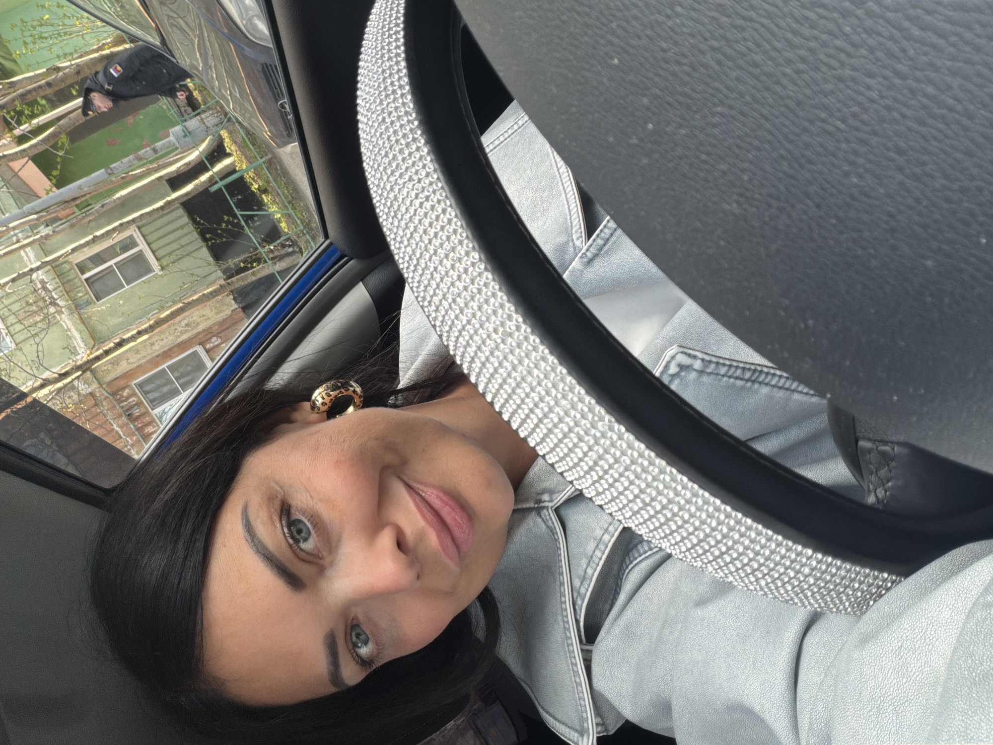 A woman with dark hair and blue eyes is inside a car, her face visible behind a steering wheel covered with rhinestones. She is wearing a light-colored denim jacket and a leopard-print earring. Through the car window, buildings are visible, suggesting an urban environment. Based on the location provided (Murmansk, Russia), the surrounding buildings might reflect architectural styles common in Russian cities, although the details are limited by the viewing angle. A person is vaguely visible outside of the car.