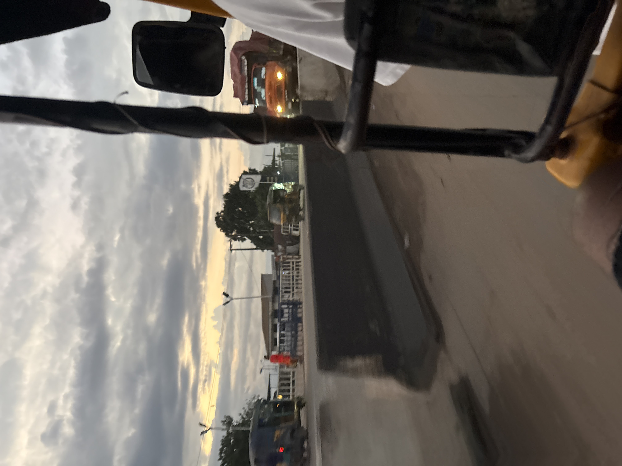 This is a street-level, eye-level, close-up photograph taken from the perspective of someone in a motorized rickshaw (also known as an 'Okada' or 'Keke') traveling on a bridge in Onitsha, Nigeria. The sky is overcast with dramatic clouds, illuminated by the setting sun, casting a soft glow. A dark barrier runs along the side of the road, suggesting the edge of the bridge. In the distance, a large orange truck with its headlights on is visible, and a yellow motorized rickshaw is passing by on the other side of the barrier. A person in a bright orange shirt is standing near a white railing. The road surface appears wet, possibly from recent rain. The overall impression is one of a busy urban environment during a transitional time of day, with a focus on the movement and vehicles characteristic of Onitsha.