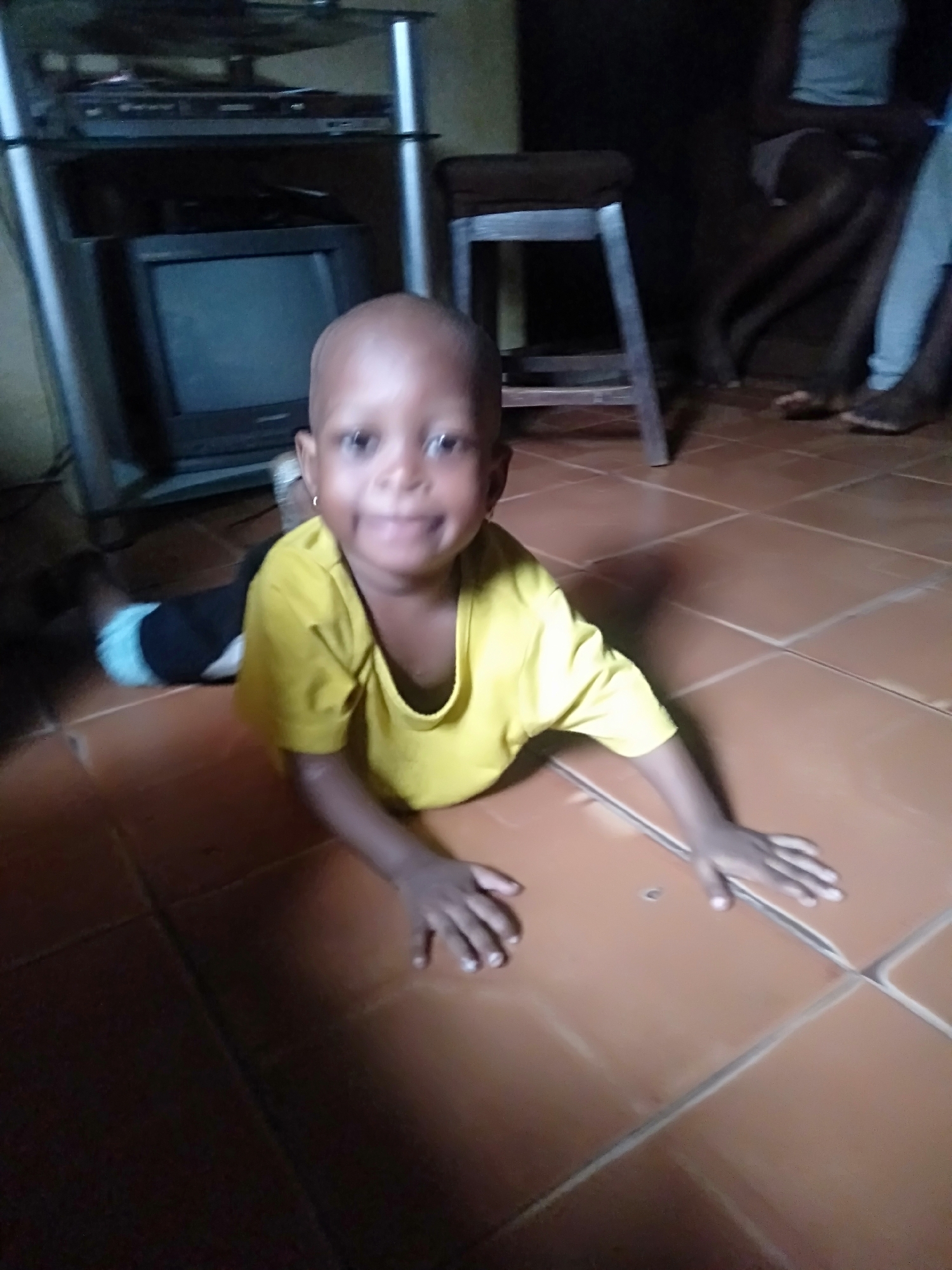 A young child, appearing to be a toddler, is the central subject of the image, lying on their stomach on a tiled floor. The child has very short hair or is bald, and is wearing a bright yellow short-sleeved t-shirt. They are looking directly at the camera with a wide, happy smile, their arms extended forward on the floor. Small hoop earrings are visible on their ears. One of the child's legs is visible to the left, wearing a black garment with a light-colored band near the ankle.
The setting is an indoor room, likely a living area, characterized by a floor made of large, square, light brown or terracotta-colored tiles with visible grout lines. In the background to the left, there's a metallic and glass television stand holding an older model CRT television and other electronic devices, possibly a DVD player or similar, stacked above it. To the right of the child, a wooden stool is visible. Further in the background on the right, the legs and feet of at least two other individuals, dressed in light-colored clothing, can be seen, suggesting the presence of more people in the room.
The overall lighting in the room is dim, casting shadows, particularly in the background areas. The child's playful posture and bright smile convey a cheerful and joyful emotion. There is no visible text in the image.