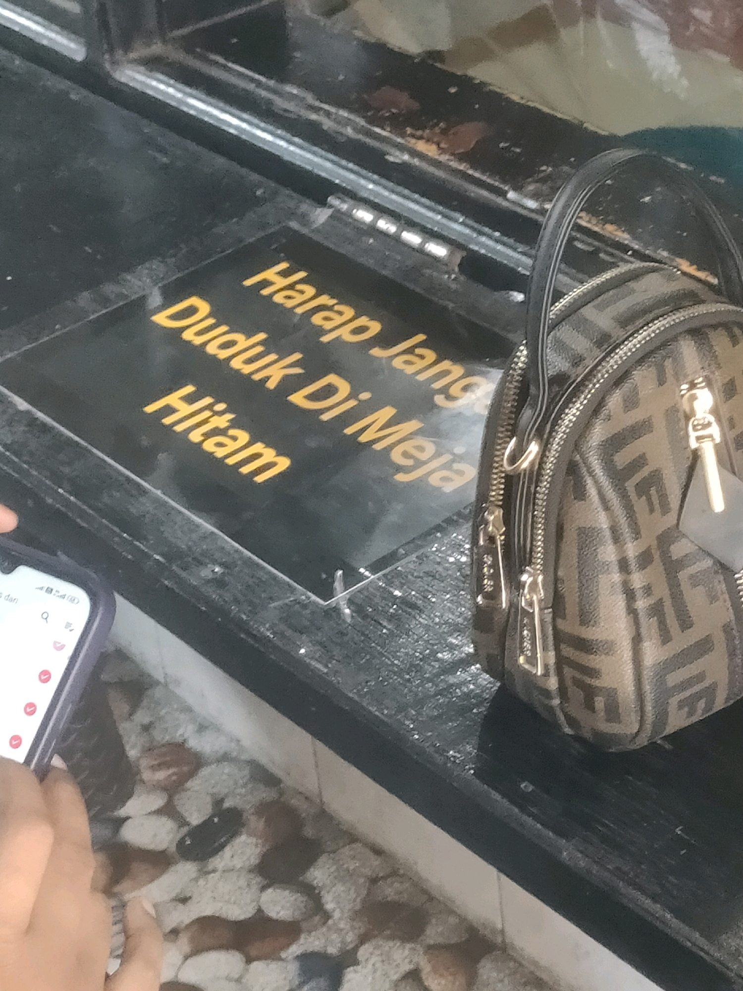 A close-up shot shows a black surface with yellow text on it, reading "Harap Duduk Di Meja Hitam." In the background, a small, brown and black patterned handbag with gold zippers is visible. A hand holding a smartphone is in the lower-left corner. The scene appears to be outdoors, possibly a street stall or restaurant in Surabaya, Indonesia. The ground below is covered in small, multicolored stones.