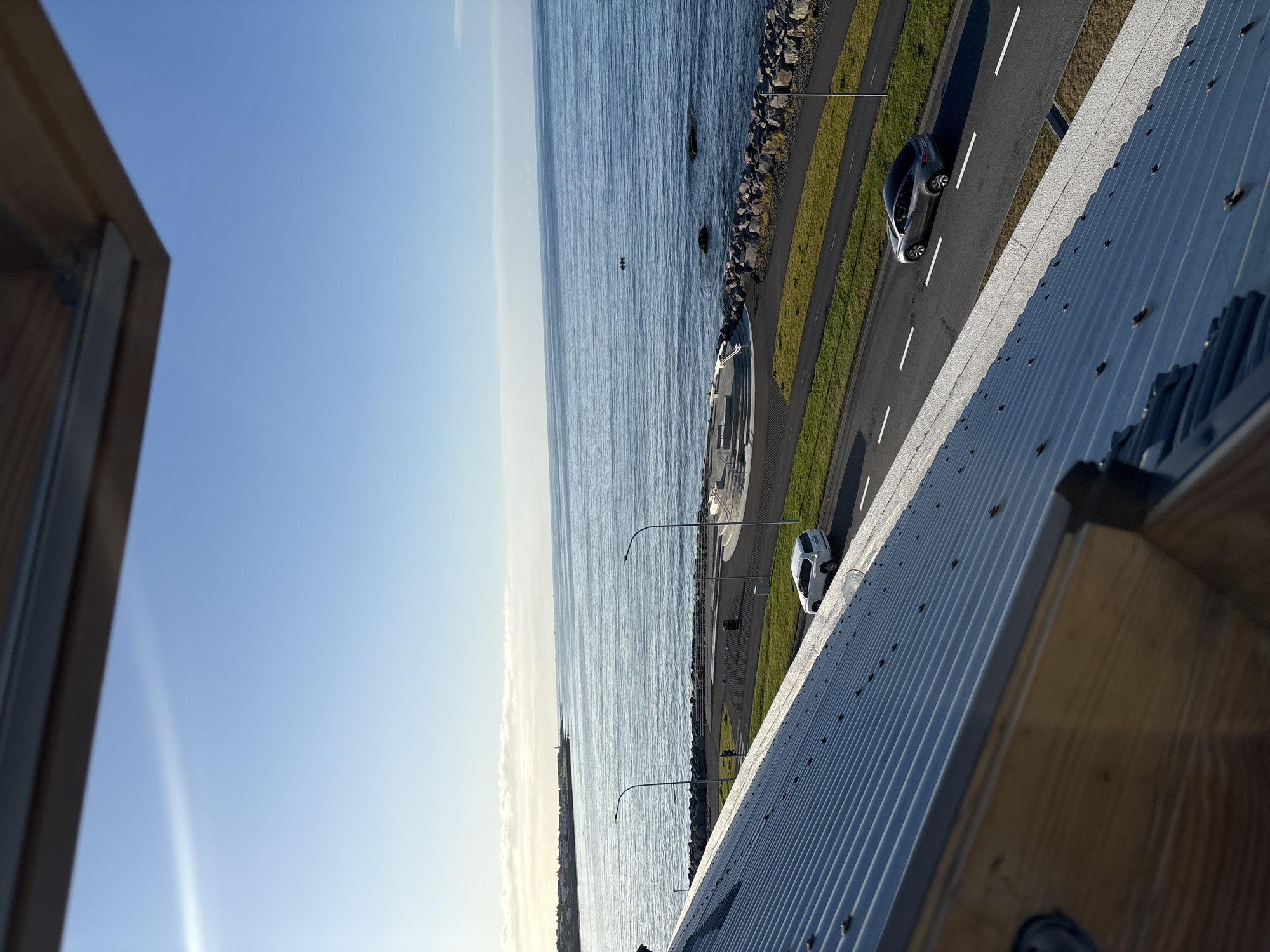 This image captures a scene in Reykjavik, Iceland, likely taken from a rooftop overlooking the waterfront. In the foreground, the ribbed metal roof of a building dominates the lower portion of the frame, with wooden beams visible at the very bottom edge. Beyond the roof, a paved road runs parallel to the ocean. A white SUV is parked on the side of the road, and a silver sedan is driving on the road. The ocean stretches to the horizon, with a calm, blue surface reflecting the clear sky. In the distance, a shoreline with buildings and a lighthouse can be seen. A small, circular seating area with steps is situated on a paved promenade along the water's edge, bordered by a rocky breakwater. Two figures in a small boat are visible on the water. The overall impression is one of a peaceful, sunny day in a coastal city.