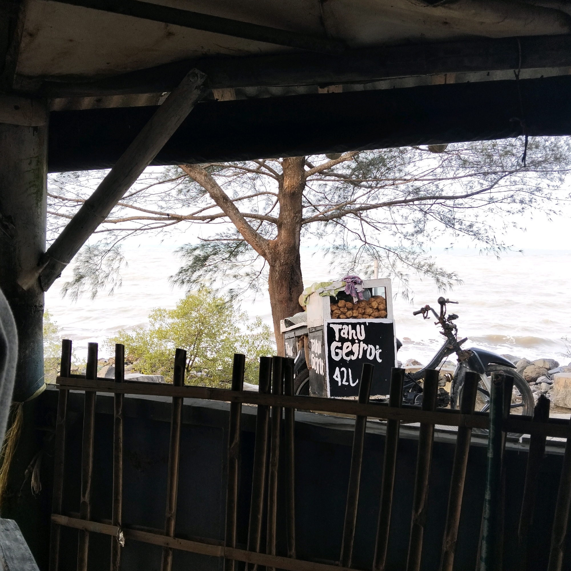 The image depicts a scene at the edge of a body of water, possibly a beach or a large lake, with a weathered wooden structure in the foreground. A food cart, labeled "TAHU GEJROT 42L", is positioned outdoors near a tree. The cart is a white box with a black chalkboard displaying the text. Beside the cart is a motorcycle with its front wheel removed. The water in the background is choppy, suggesting wind or waves. The overall atmosphere is rustic and somewhat neglected. The scene is likely located in a coastal area or near a large body of water within Bandung City, Indonesia, given the mention of "TAHU GEJROT", a popular Indonesian street food originating from Cirebon, West Java, a region adjacent to Bandung.