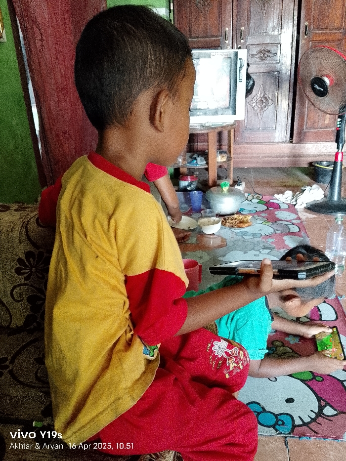 Two young children, one wearing a yellow shirt with red trim and red pants, and one wearing a green shirt, are seated on a floor covered with a colorful patterned rug. The child in the yellow shirt is holding a black tablet and looking at it. The child in the green shirt is lying on the floor with his face turned away from the camera, and looking at a smartphone. The scene appears to be inside a home in Anjatan, Indonesia. The home features wooden cabinets, a television, and a fan. There is a small bowl of food and a cup on the floor, and a few other items of furniture are visible in the background. The image was taken with a smartphone camera and the date and time are recorded as "16 Apr 2023, 10.51".