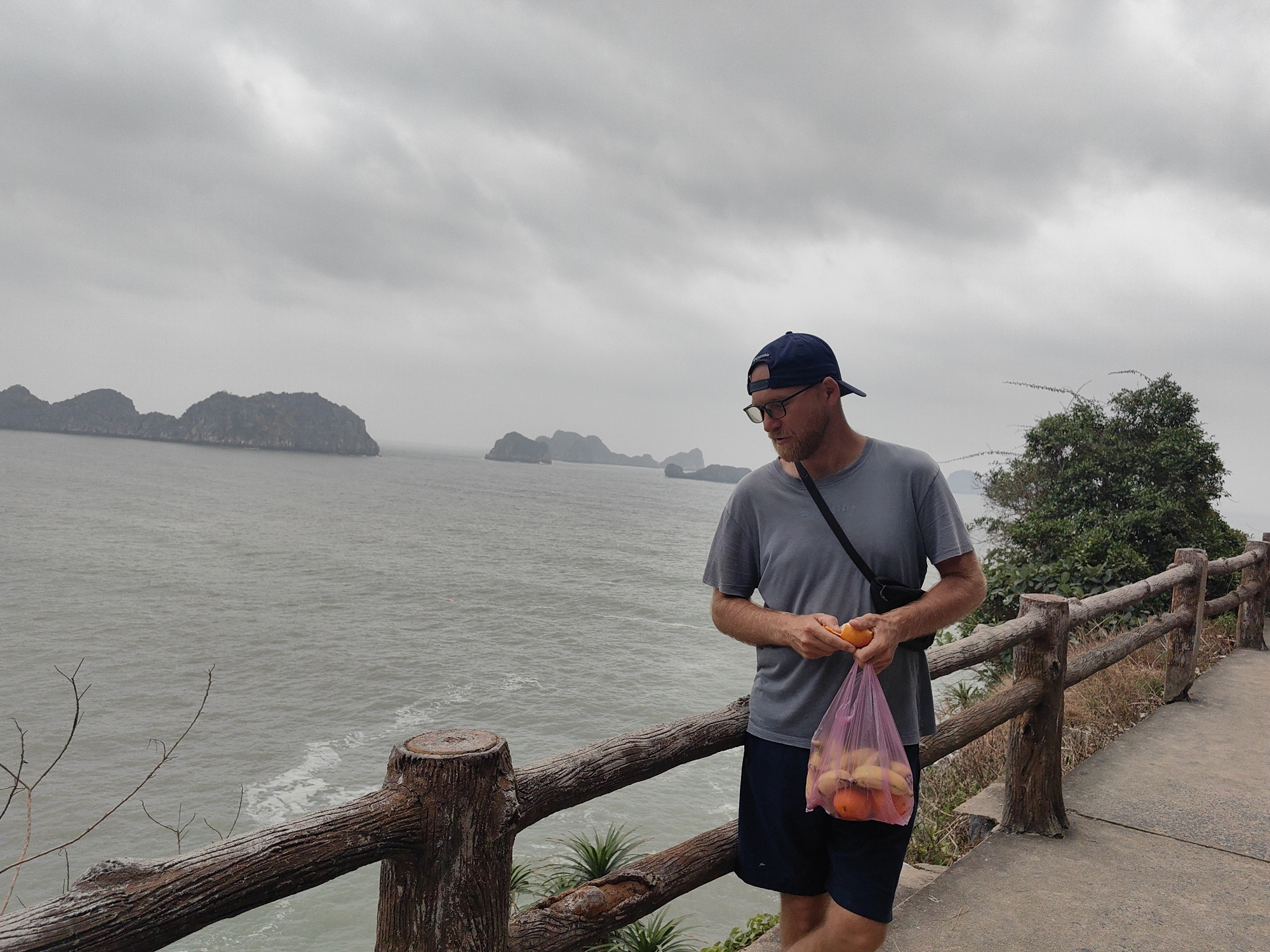 A man in a gray t-shirt, dark blue shorts, a dark blue baseball cap, and glasses stands next to a wooden railing holding a plastic bag of fruit. He is peeling an orange. A shoulder bag strap is visible across his torso. The railing runs along a paved path. Beyond the railing is a body of water with islands in the distance. The sky is overcast. A tree is to the man's right. The location is in the city of Hải Phòng, Vietnam.
