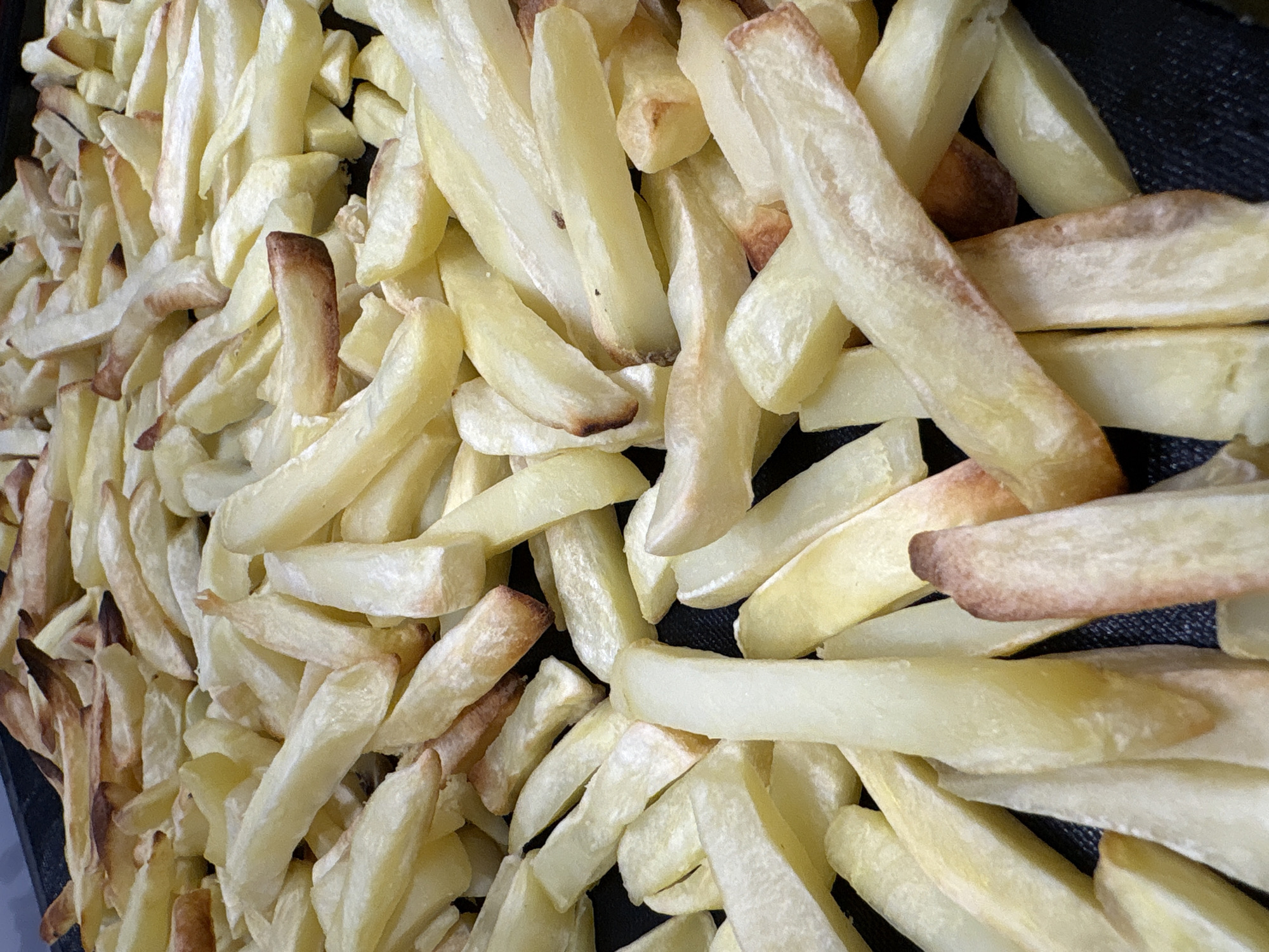 The image shows a close-up of a heap of pale yellow french fries with some edges and tips slightly browned. They are piled on a dark gray or black surface. There is no indication of the city of Izhevsk, Russia or any identifying information about the location, people, or interactions in the image.