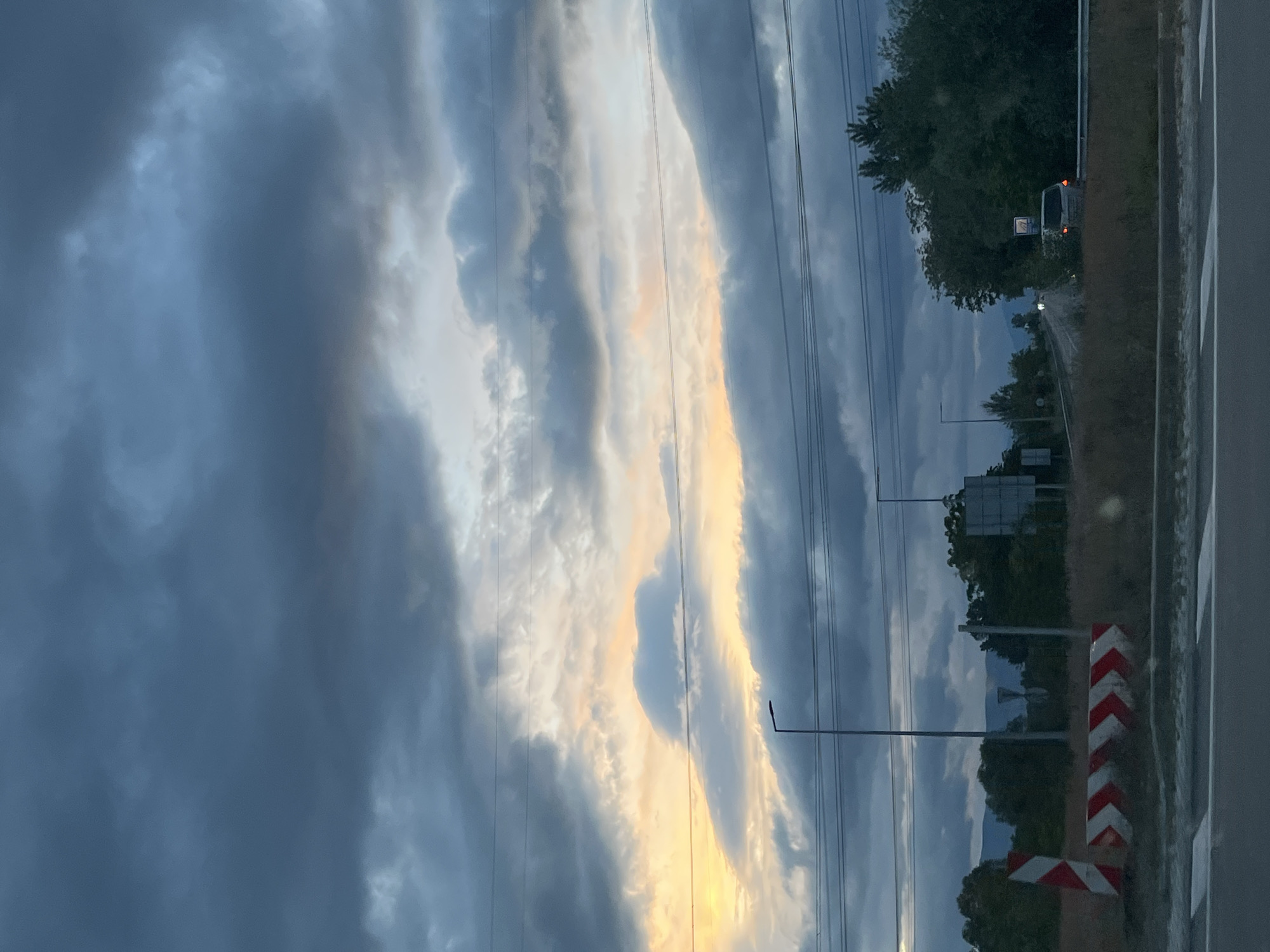 The image depicts an overcast sky with streaks of light visible through the clouds, taken from beside a road in Blagoevgrad, Bulgaria. The roadside features a guardrail with red and white chevron markings. Power lines run along the side of the road. Vegetation, including trees, is present on the shoulder of the road. A vehicle, likely a van, is visible in the distance, with a traffic sign visible nearby. A street light is also present.