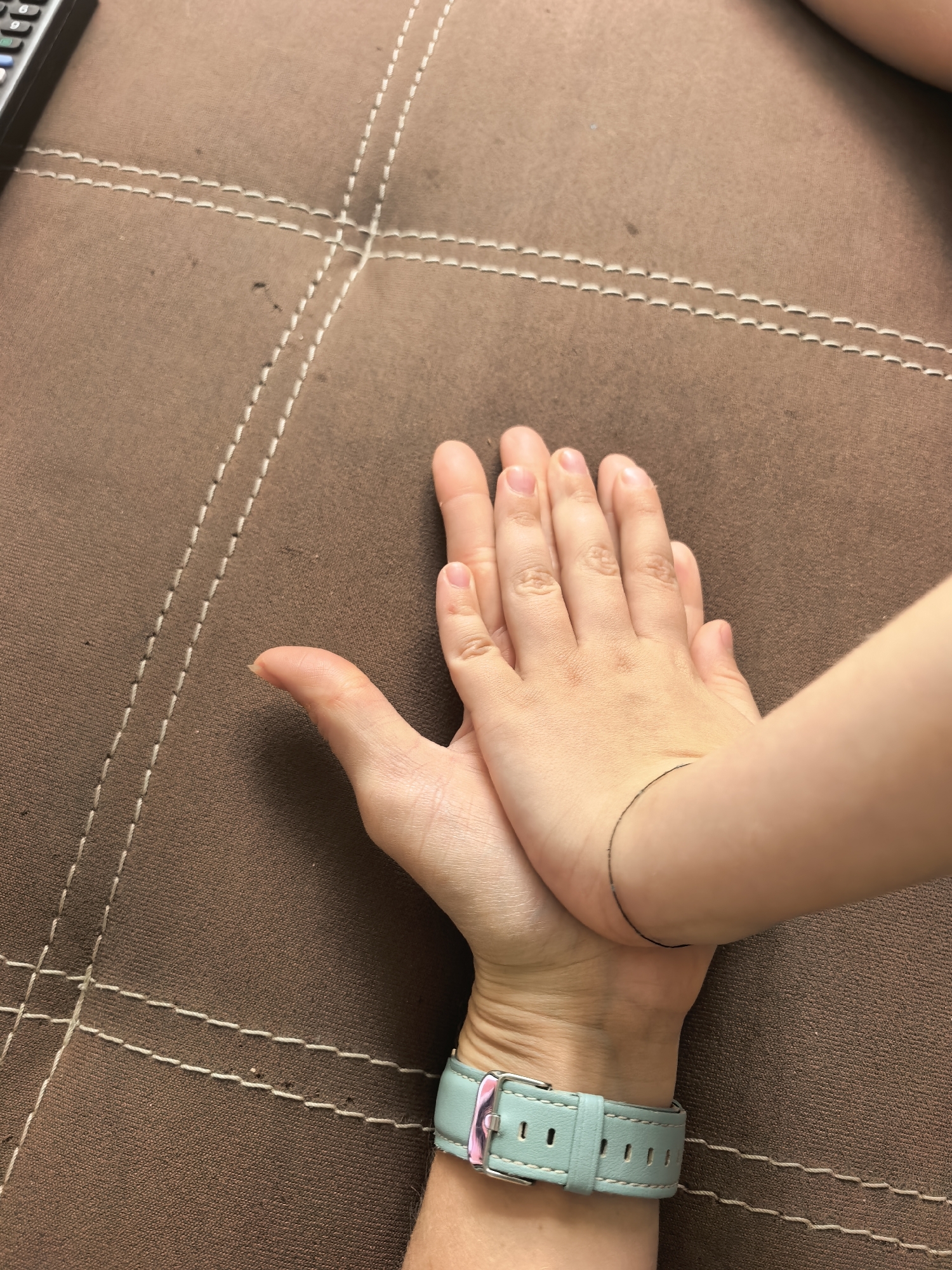 An overhead medium shot shows two hands, one adult and one child, placed palm-to-palm on a brown textured surface, likely a couch. The adult's hand is larger and positioned below the child's hand. The child's hand is resting on the adult's hand, with fingers slightly spread. A light blue watch with a silver buckle is visible on the adult's wrist. The brown surface has visible white stitching that forms a diamond pattern. The image does not provide any information about the location in the city of Sterlitamak, Russia.