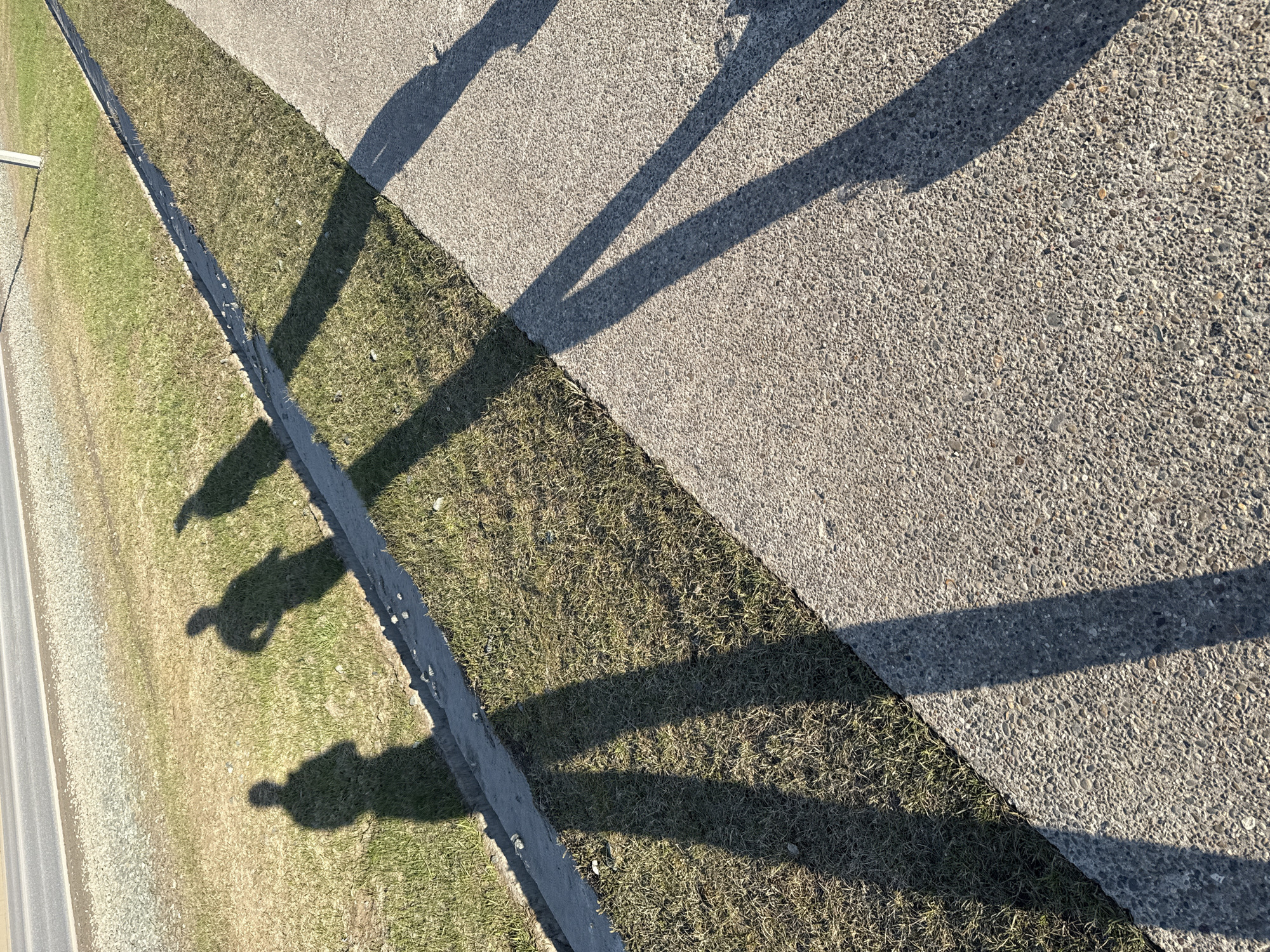 The image depicts a gray concrete surface that transitions to a grassy area. A narrow, paved road runs adjacent to the grass. Shadows, seemingly cast by a metal structure, stretch across the concrete and grass. Three distinct human-like shadows are visible on the grassy area. Based on the location, this is likely a public space in Chaikovsky, Russia, possibly a park, roadside, or pedestrian area. The image captures shadows suggesting the presence of structures and people.