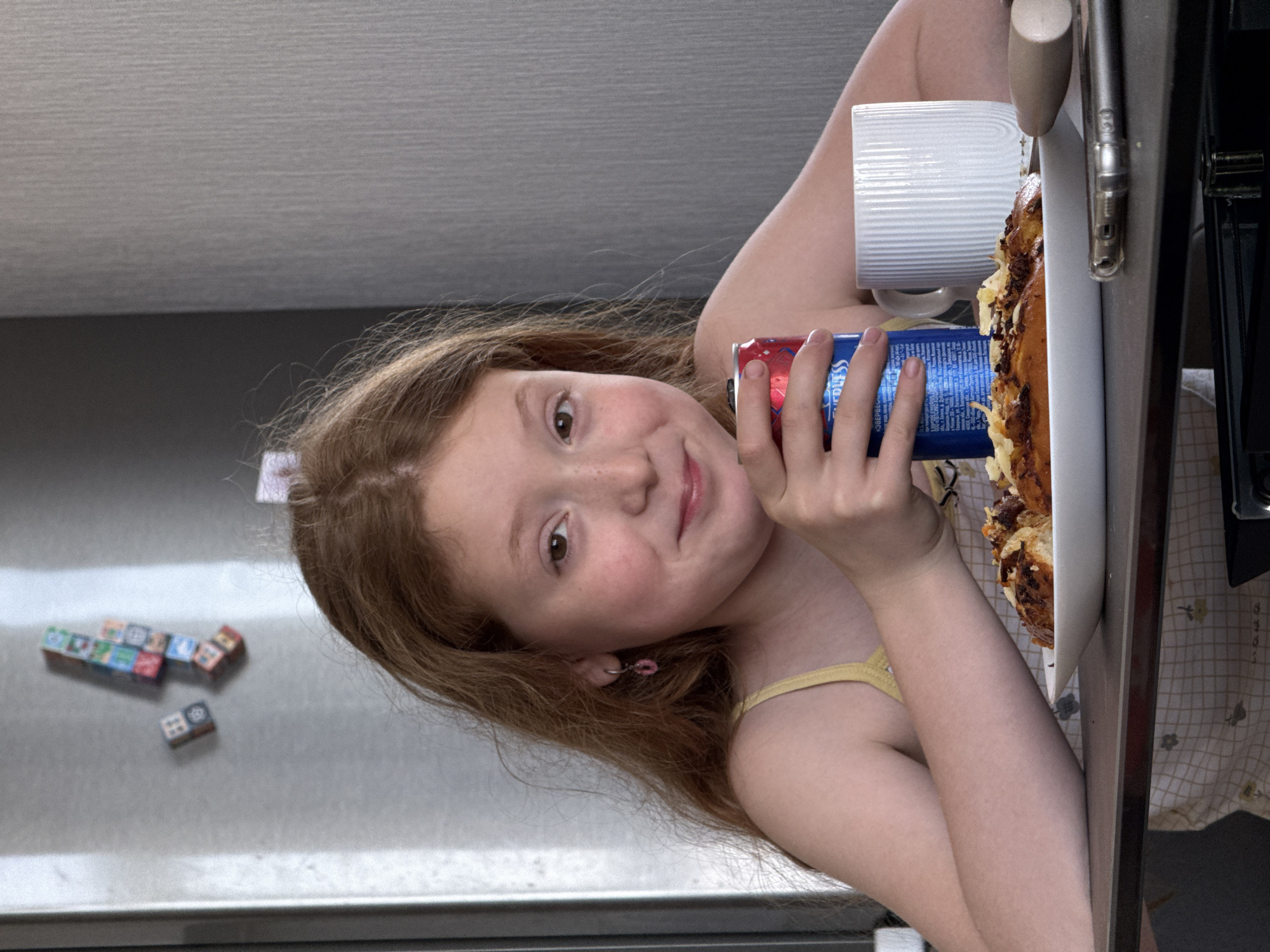 A young girl with reddish hair, light skin, and freckles is leaning towards the camera holding a can of soda in her right hand. She's wearing a yellow tank top. Beside her is a white mug and a plate of food that appears to be some kind of baked dish. The setting appears to be a kitchen, with a stainless-steel refrigerator visible behind her that has small square magnets on it. The background includes a beige ceiling. There's no information to ascertain that the location is Rostov-on-Don, Russia.
