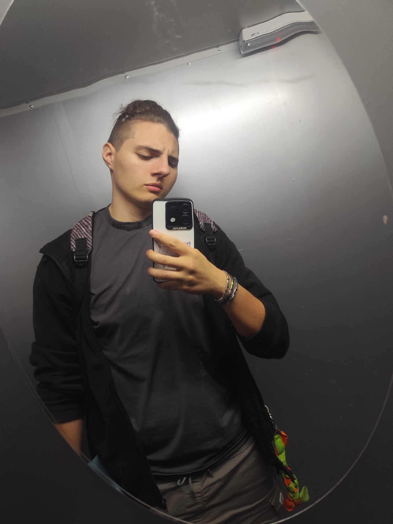 In a dimly lit elevator with gray metallic walls, a young person with a man bun hairstyle takes a selfie in a large, slightly convex mirror. They are wearing a dark zip-up jacket over a dark grey t-shirt, and khaki-colored pants. A backpack with a checkered strap is visible on their left shoulder, and a small keychain with green and orange figures hangs from their right side. The elevator ceiling features a white emergency light with a red indicator. The overall scene suggests a mundane moment captured during transit within a building, possibly in an urban environment like Sofia, Bulgaria, though no specific landmarks or indicators of the city are present in the image.