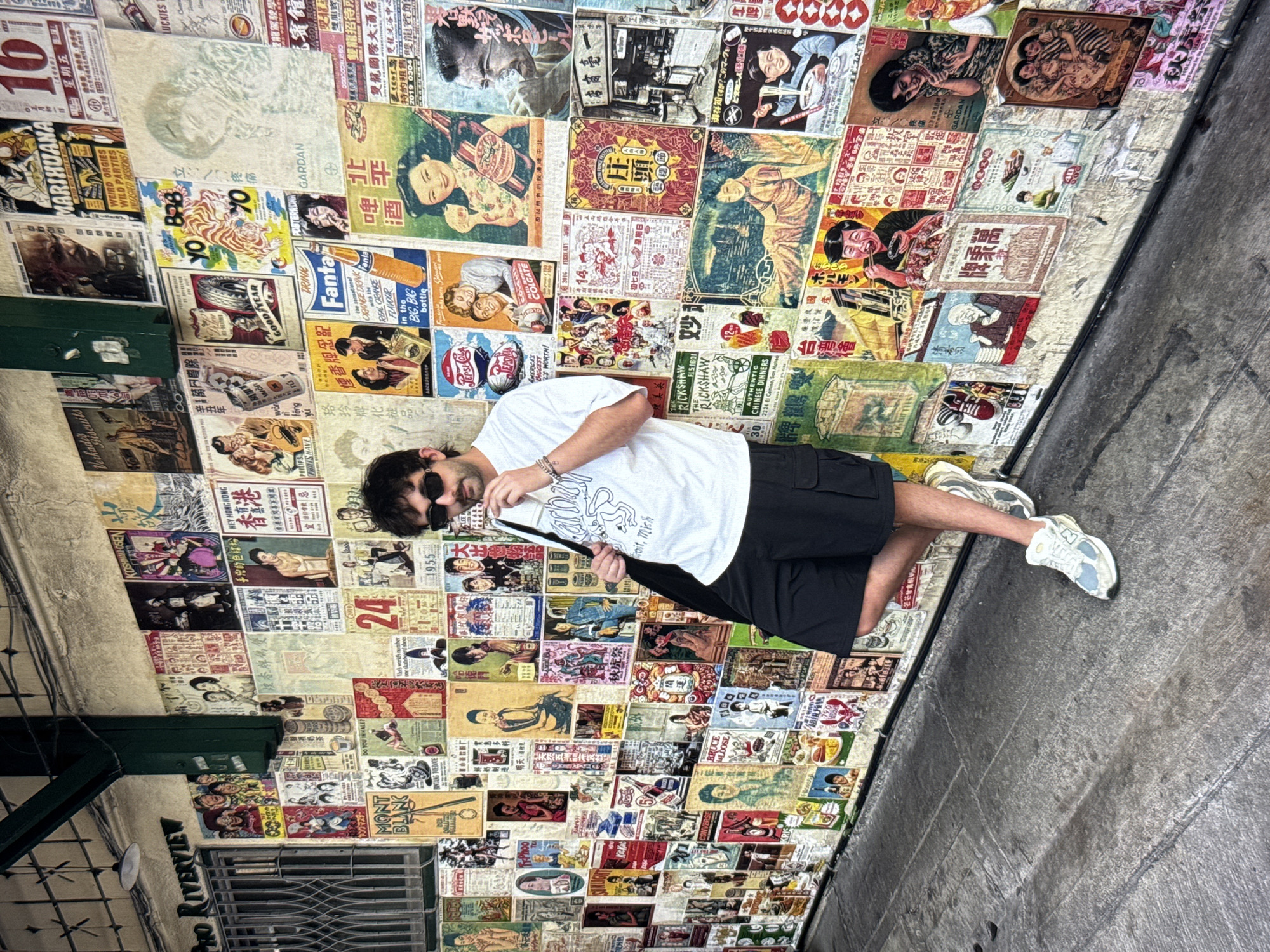 The image shows a man leaning against a wall covered in vintage posters. The man is wearing a white t-shirt, black shorts, and white sneakers. He has dark hair and is wearing sunglasses. The posters on the wall are colorful and varied, featuring advertisements, illustrations, and text in what appears to be Chinese and English. Some posters depict people, products like Fanta and Pepsi-Cola, and various scenes. The wall itself is rough and textured, suggesting an outdoor urban environment. The ground in front of the wall is a weathered, grey paved surface. The overall impression is of a visually rich, possibly historic, street scene. This location is not in Moscow, Russia; it appears to be an Asian city given the language on the posters.