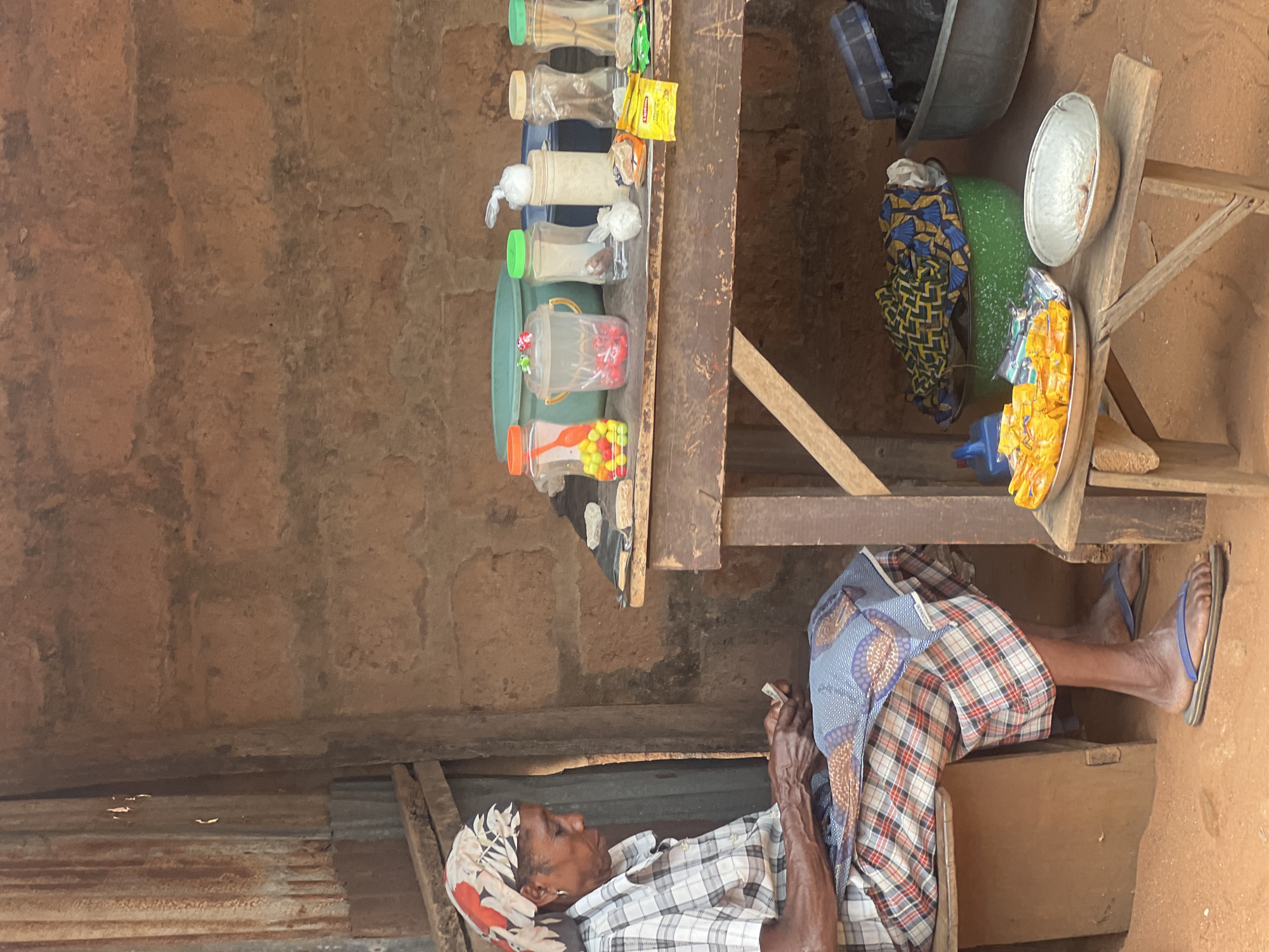 A woman sits in front of a stall in an unknown location. The stall features a wooden shelf with various items for sale, including bottled candies and small packaged goods. The woman is wearing a checkered shirt, a printed headscarf, and is holding something in her hands. A variety of containers and cloths sit to the right of the stall on a wooden stool. The surrounding environment includes what appears to be a mud-walled structure.