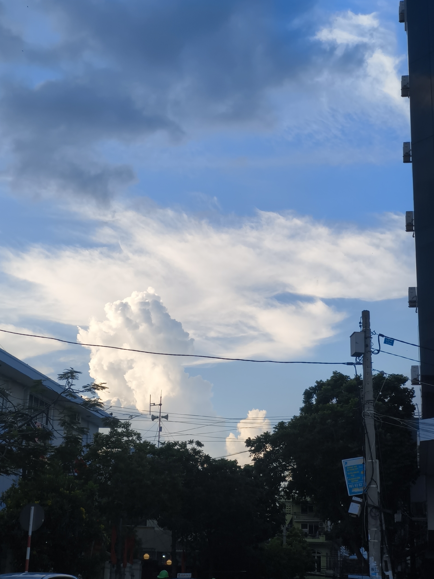 The image captures a view of the sky and urban landscape in Bac Nha Trang Ward, Vietnam. The sky is predominantly blue with various cloud formations. On the left side of the frame, a building with a blue roof is visible, partially obscured by trees. A traffic sign with red and white stripes is positioned near the building. In the center and right of the image, a utility pole stands tall, adorned with electrical wires, boxes, and a blue sign. The pole is surrounded by lush green trees. Further in the background, another building with a pale green facade is discernible. A sliver of a dark-colored building is present on the far right edge of the image. The scene is illuminated by natural light, suggesting it was taken during the daytime.