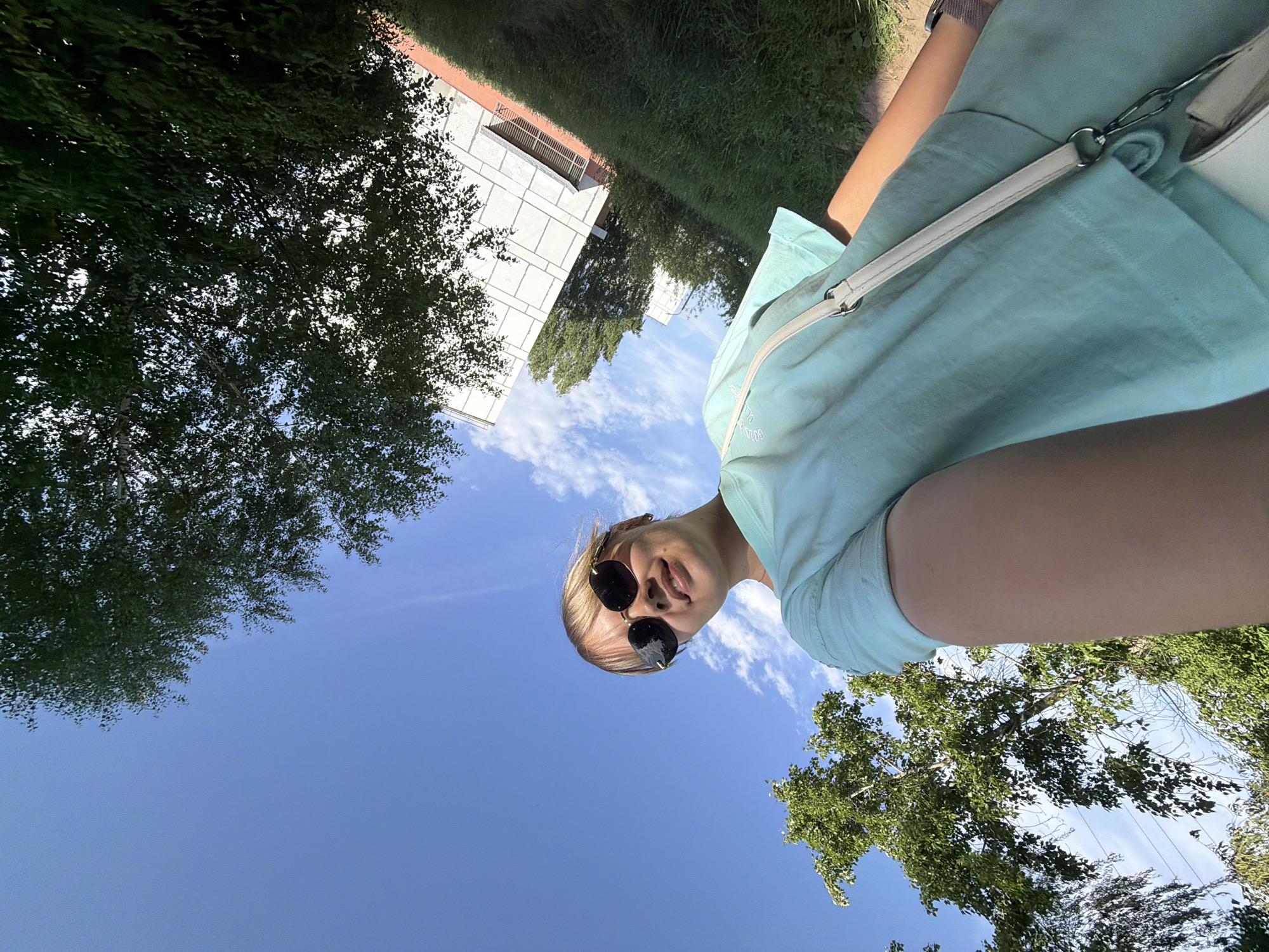 A low-angle photograph shows a person with light skin and blonde hair wearing sunglasses and a light blue shirt. A white shoulder bag is slung across their body. The background features a clear blue sky with scattered clouds, tree foliage on the left and right sides of the frame, and a modern building with a white facade in the upper-left quadrant. The location is Chaikovsky, Russia, based on the style of architecture and vegetation typically found in the region. The person is likely taking a selfie or someone is taking a picture of the person from a low angle.