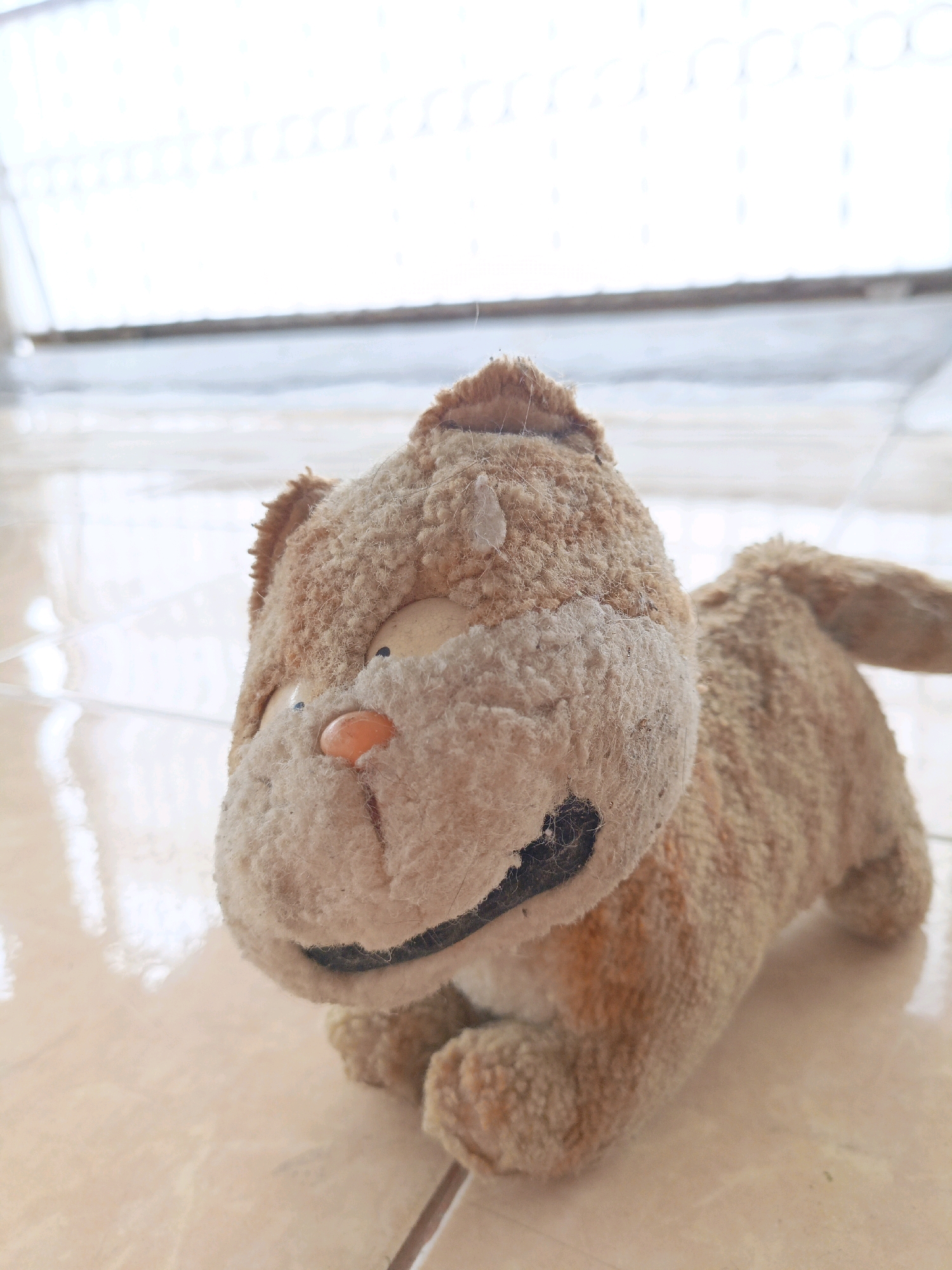 A close-up, eye-level shot shows a worn, light brown stuffed cat toy lying on a light brown tiled floor. The toy is positioned to the right of the frame, facing left. Its fur is matted and appears dusty, with some darker brown patches. The cat has large, round, light-colored eyes, a small orange nose, and a black stitched mouth. Its ears are small and rounded. In the background, a blurred white light suggests a window or screen door with horizontal elements. The floor tiles have a subtle sheen, reflecting the light. The scene appears to be indoors, possibly in a home, in the city of Medan, Indonesia. There are no people or animals present other than the stuffed toy.