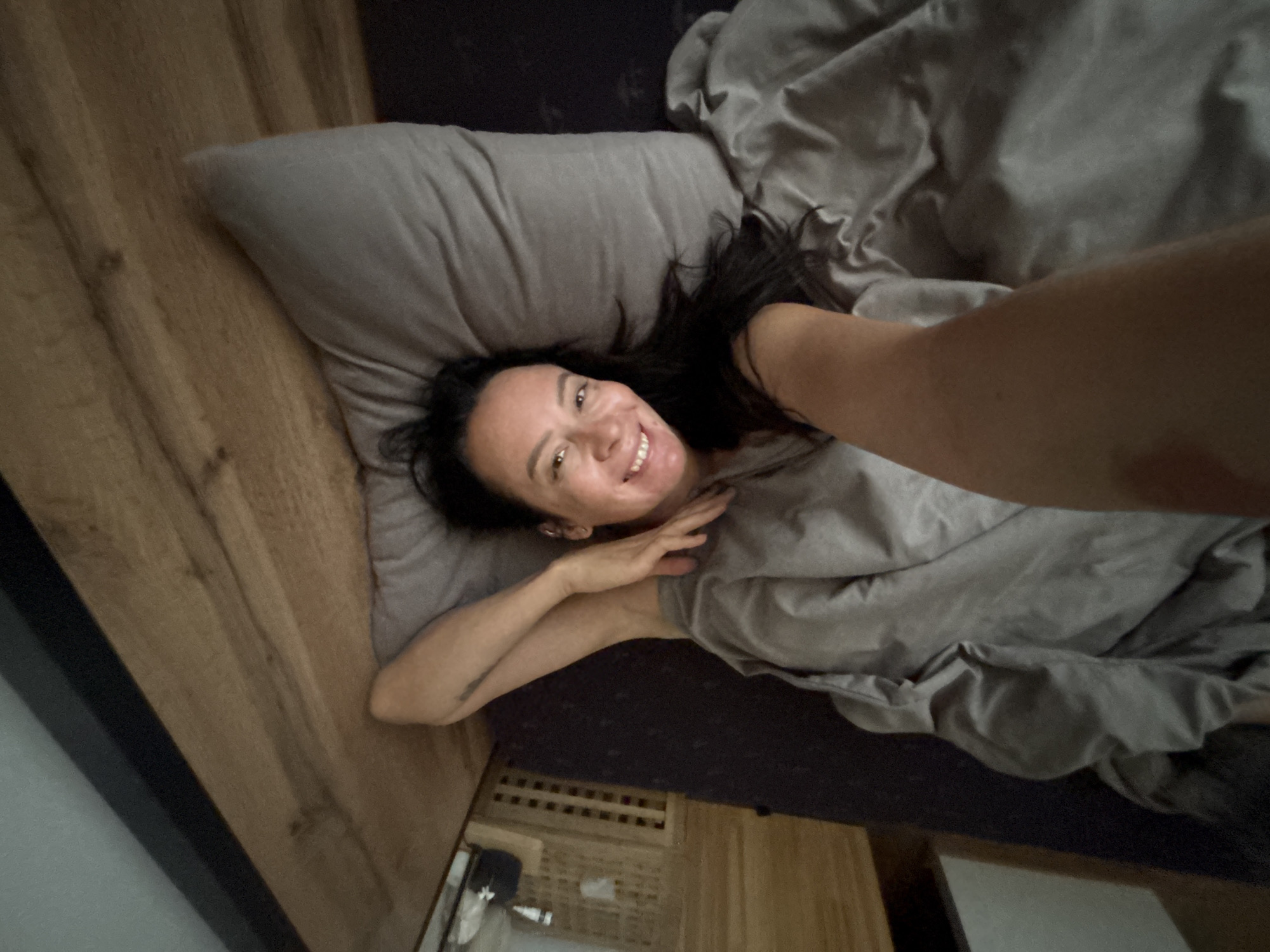 A woman is lying in bed in Chaikovsky, Russia. She is facing the camera with her head resting on a gray pillow, covered with gray sheets. She is smiling and holding up her arm to take a selfie. The bed is against a wooden headboard. A nightstand is visible beneath the bed.