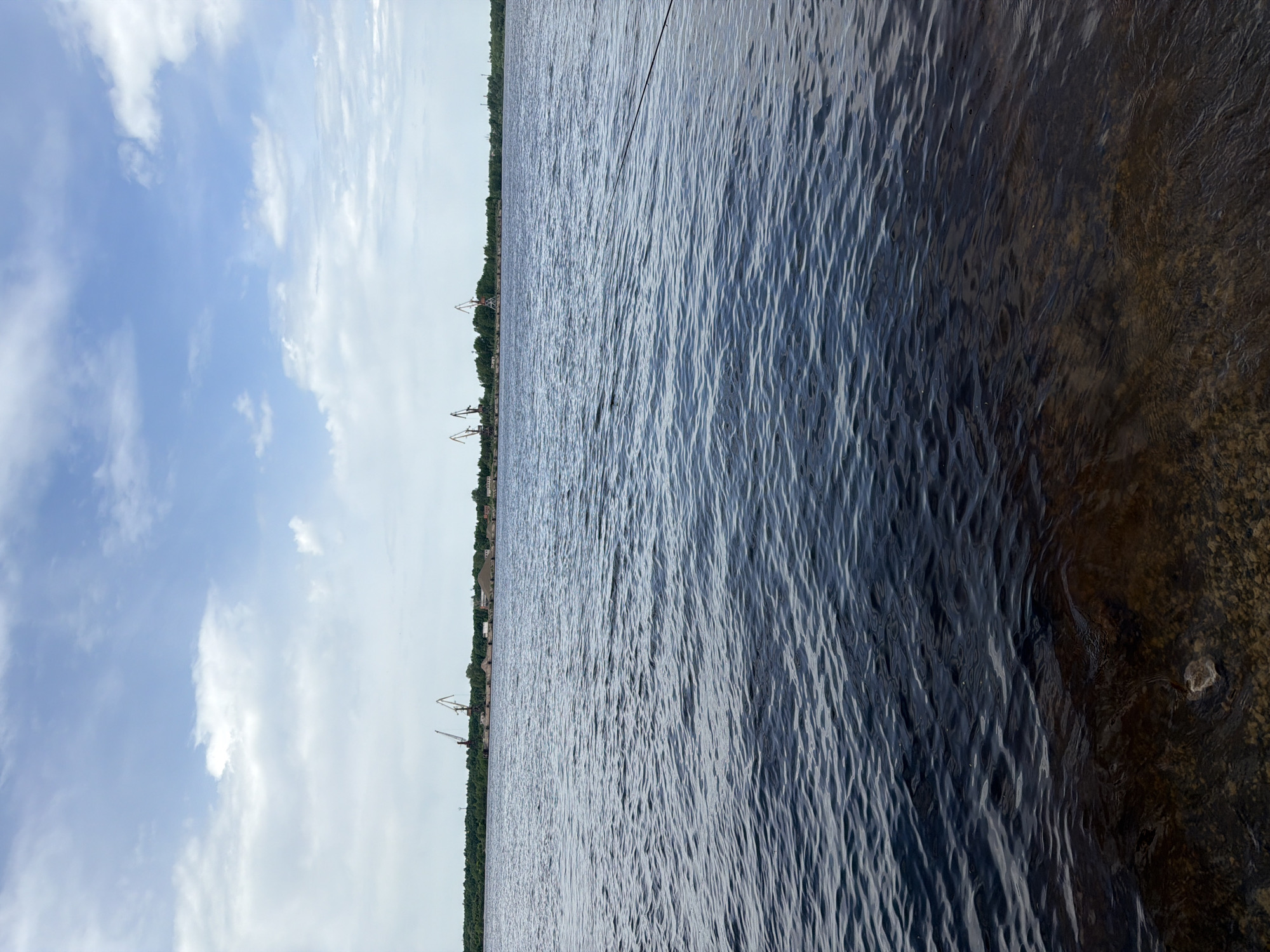 The image shows a body of water, likely a river or lake, with a visible shoreline in the distance. The water is dark and rippled, suggesting a breeze. The shoreline is lined with trees and buildings, including several cranes, indicating a port or industrial area. The sky is partially cloudy with a mix of blue sky and white clouds. The right side of the image reveals the edge of the body of water with a view of the bank.
Given the presence of cranes and the general appearance, the location may be Noviy Urengoy in Russia, as the prompt suggests.