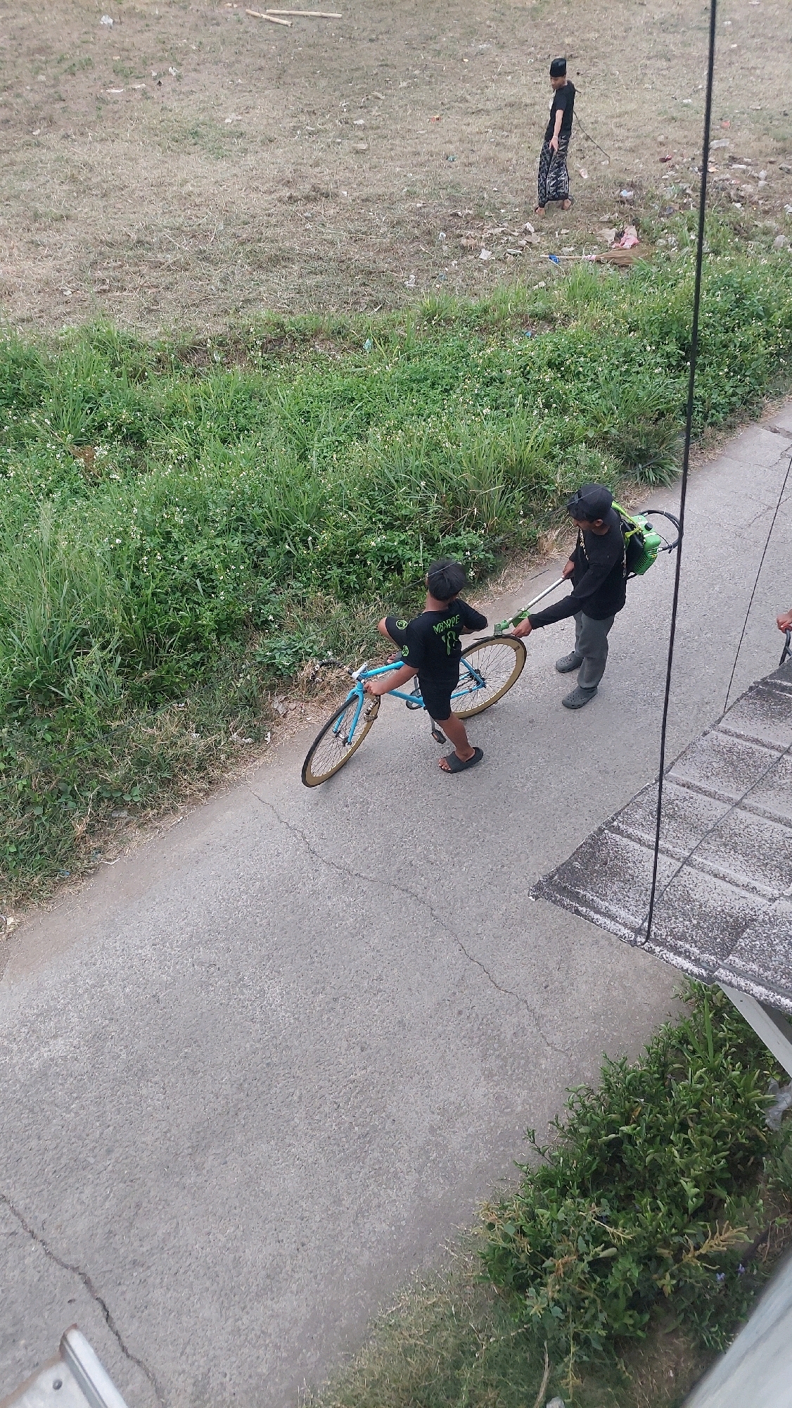 In Bojong Gede, Indonesia, two young males are observed on a paved path next to a grassy verge. The male on the left, wearing a black t-shirt with "VANS" printed in green on the back and black shorts, is holding a blue bicycle by the handlebars, facing away from the viewer. He is wearing black sandals. The male on the right, also wearing a black t-shirt and black cap, is carrying a green backpack-style leaf blower, with the blower's hose extended towards the first male, who appears to be operating it. This male is wearing grey trousers and dark grey footwear. In the background, a third individual, wearing a black t-shirt and patterned sarong, stands in a dry, grassy area with scattered debris. The environment suggests an outdoor, potentially semi-rural or unkempt urban setting.