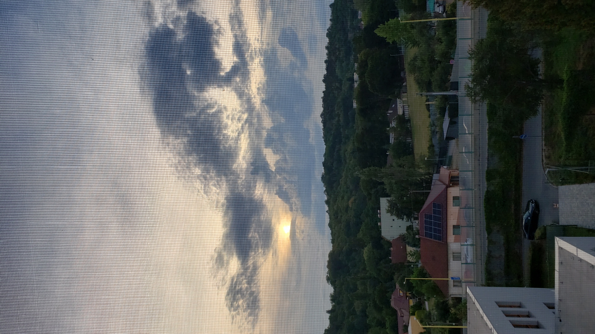 The image depicts a view through a window screen at dusk, overlooking a suburban landscape. In the distance, a hilly area covered with dense green trees forms the backdrop. The sky is filled with dramatic clouds, with a sliver of the setting sun visible through a gap, casting a warm glow. In the mid-ground, several residential buildings are visible, one of which has solar panels installed on its roof. A dark-colored car is driving on a road in the foreground. The scene suggests a peaceful evening in a residential area, likely on the outskirts of a city given the combination of housing and natural landscape. Based on the architectural style and the presence of solar panels, the location appears to be a modern residential area. Without further context or recognizable landmarks, pinpointing the exact location within Prešov, Slovakia is not possible from the image alone.