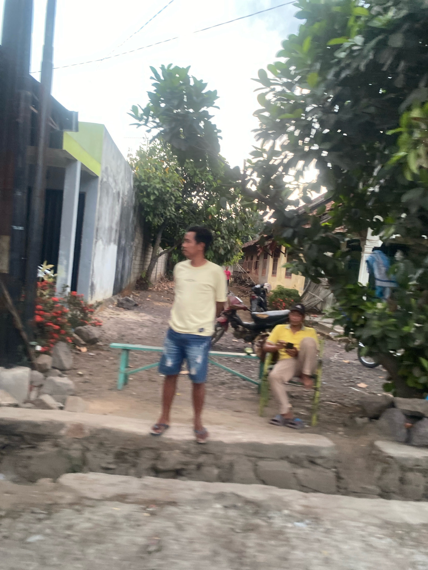 In Banjar Sari, Indonesia, a man stands on a raised concrete platform to the left of center, looking to the right. He wears a light yellow t-shirt and distressed denim shorts. To his right, a green metal bench is visible. Further right, a second man sits on a green metal chair, looking down at a smartphone in his hands. He wears a yellow t-shirt and light brown pants. The background features a building with light yellow and gray walls, trees with lush green foliage, and a dirt ground with scattered rocks. Several motorcycles are parked in the middle ground. The overall lighting suggests it is daytime with some overcast.