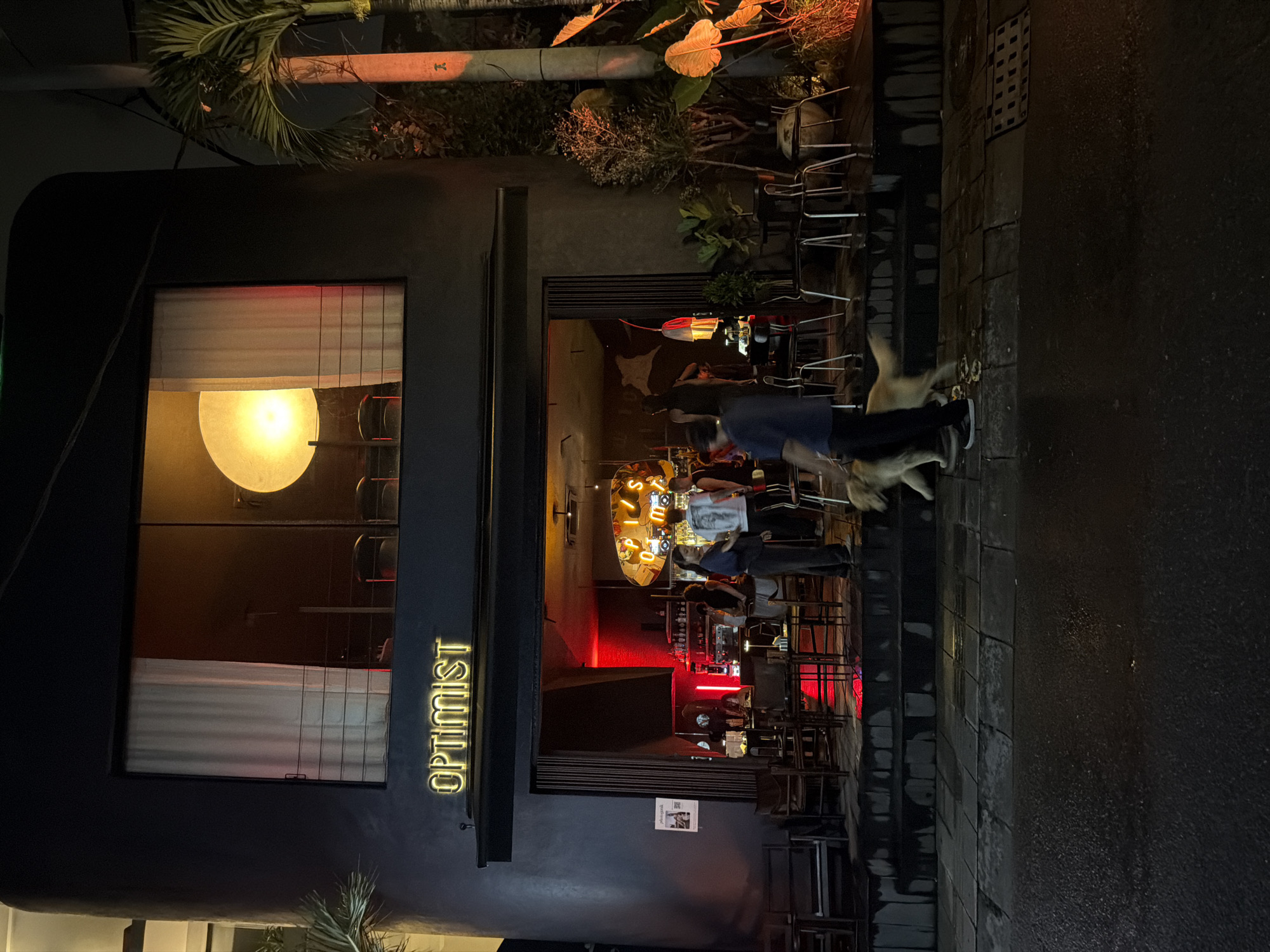 The image depicts a street view at night outside a building with the neon sign "OPTIMIST" in yellow lettering. The building appears to be a restaurant or bar, with large windows revealing a warmly lit interior. Inside, several people are visible, some standing and conversing, and others seated. The interior is decorated with red lighting and a prominent circular mirror with the word "OPTIMIST" also reflected on it.
Outside, there are several chairs and tables arranged on the sidewalk, suggesting an outdoor seating area. Two dogs are also present on the sidewalk, one appearing to be walking alongside a person. The ground is wet, indicating recent rain or a damp evening. The scene is illuminated by the building's interior lights and the neon sign, casting a moody glow on the street. The overall atmosphere appears relaxed and urban, with a hint of the establishment's inviting ambiance. The context provided suggests this scene is located in Mengwi, Indonesia.