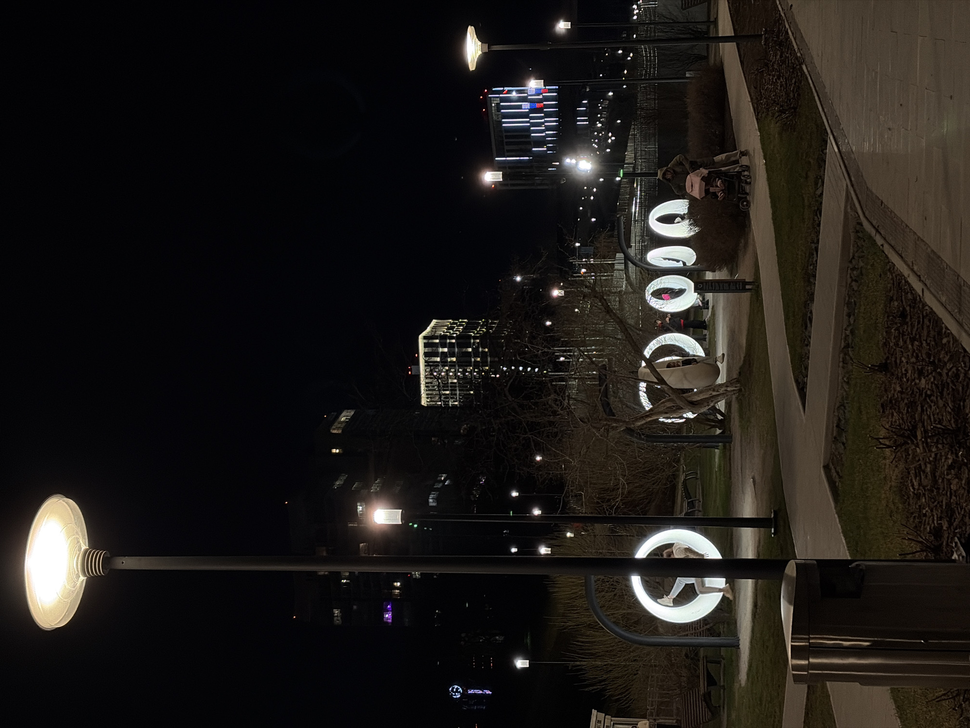 The image displays a sideways view of an urban park or pedestrian area at night. A wide, light-colored paved pathway curves through the scene, flanked by grassy sections and trees with bare branches, suggesting a colder season.
Several large, white, glowing ring-shaped installations are prominent along the pathway. These illuminated structures appear to be interactive art or play elements; a person, possibly a child, is visible inside one of them. Further along the path, an adult figure is seen pushing a baby stroller with a pink canopy. Other indistinct figures are present in the distance.
The area is well-lit by modern streetlights featuring flat, circular lamps that emit a bright white light. A large streetlight is clearly visible on the left side of the foreground. In the background, contemporary high-rise buildings stand against the dark sky, their numerous windows brightly illuminated, creating striking patterns of light characteristic of an urban cityscape. A metallic, cylindrical trash bin is partially visible in the foreground on the right. The scene captures a lively yet serene evening atmosphere, characteristic of an urban park, possibly in Perm, Russia. No discernible text is visible within the image.