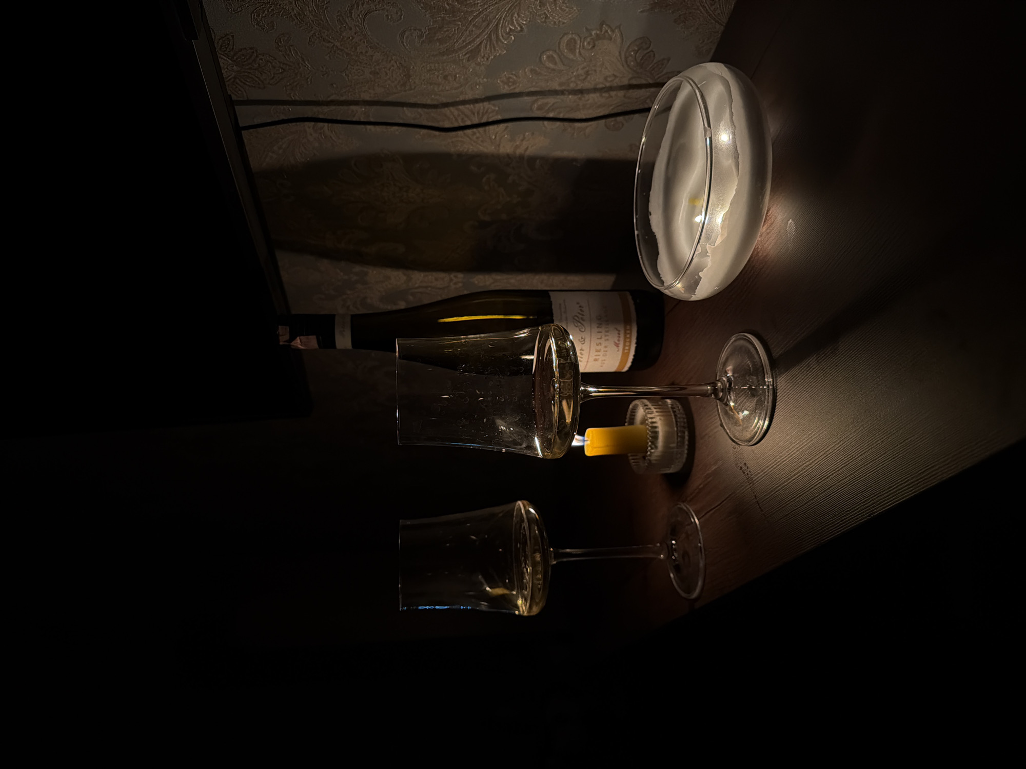 The image presents a still life scene. Two wine glasses are laid horizontally, partially visible on the lower left. A bottle of white wine is situated above them. Next to the bottle is a glass container with a candle inside. A separate small candle in a holder sits beside one of the glasses. The scene is illuminated by candlelight in a dark environment. The background features patterned wallpaper. This image may have been taken indoors, possibly a private home, in Hrodna, Belarus.