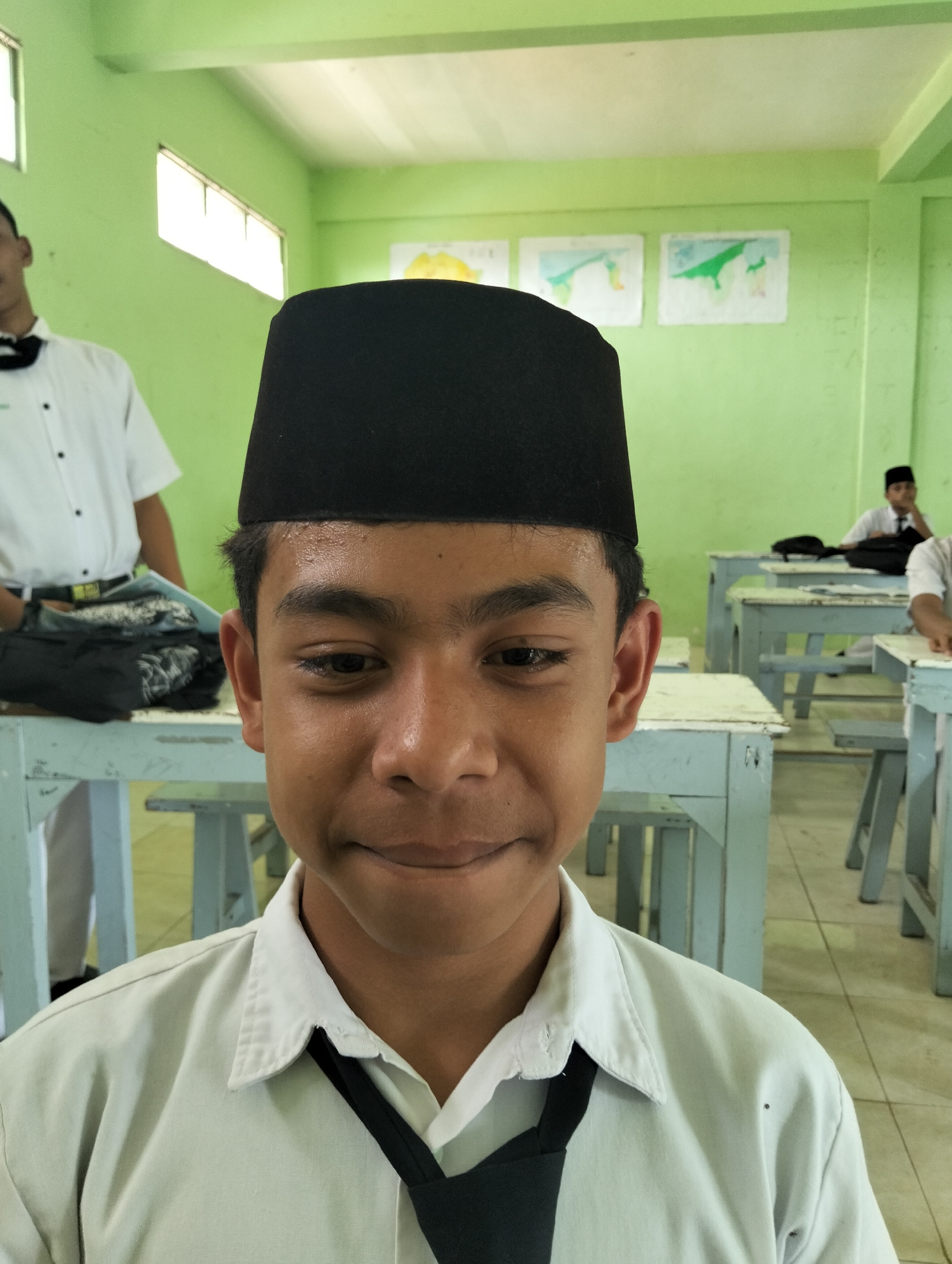 A young male wearing a white collared shirt and a black peci is looking towards the camera. The peci is worn centrally on his head. The shirt has a black tie knotted and tucked under the collar. The background is a classroom setting with green walls. Two windows are visible on the left wall, letting in bright light. On the back wall, two framed drawings or posters are displayed. Several blue wooden desks with light-colored tops are arranged in rows. In the background, other students can be seen seated at the desks, also wearing similar attire. The flooring is tiled. The scene appears to be in a school environment in Kranji, Indonesia.