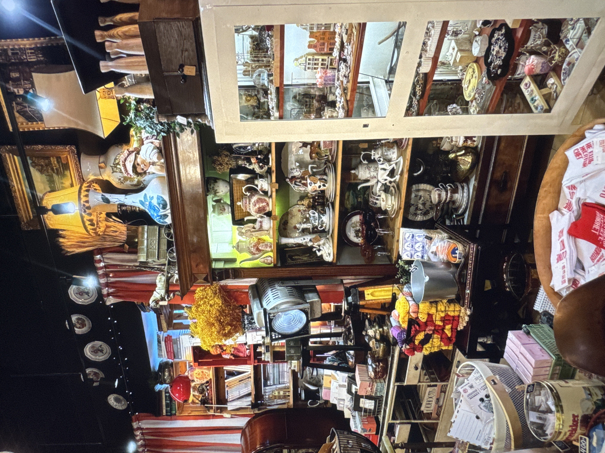 This media file captures a cluttered shop interior, possibly an antique or curio shop, filled with a diverse array of objects. The scene is densely packed with items, suggesting a place where many trinkets and collectibles are displayed.
Prominently featured are shelves and cabinets brimming with porcelain items, including teacups, saucers, and decorative vases, many adorned with intricate patterns in red and white. There are also vintage lamps with shades, and what appear to be framed artworks or prints. In one section, a collection of small, model buildings is visible, set against a backdrop that resembles a city scene. Elsewhere, decorative plates are hung in a line. A spotlight fixture is positioned above a collection of books and other items. In the foreground, a woven basket contains what looks like postcards or letters, and a large tin can is also visible. The overall impression is one of abundance and perhaps a touch of nostalgia.
Visible text on some items includes: "LOVE", "CHEF", and "Новинки". The lighting suggests an indoor setting, possibly during the day or with artificial lighting creating a warm ambiance. No people are visible in the frame, and there are no indications of weather. The presence of text in Cyrillic script, specifically "Новинки", suggests a location in Russia, consistent with the provided location context. The sheer volume and variety of items imply a rich collection of curiosities.